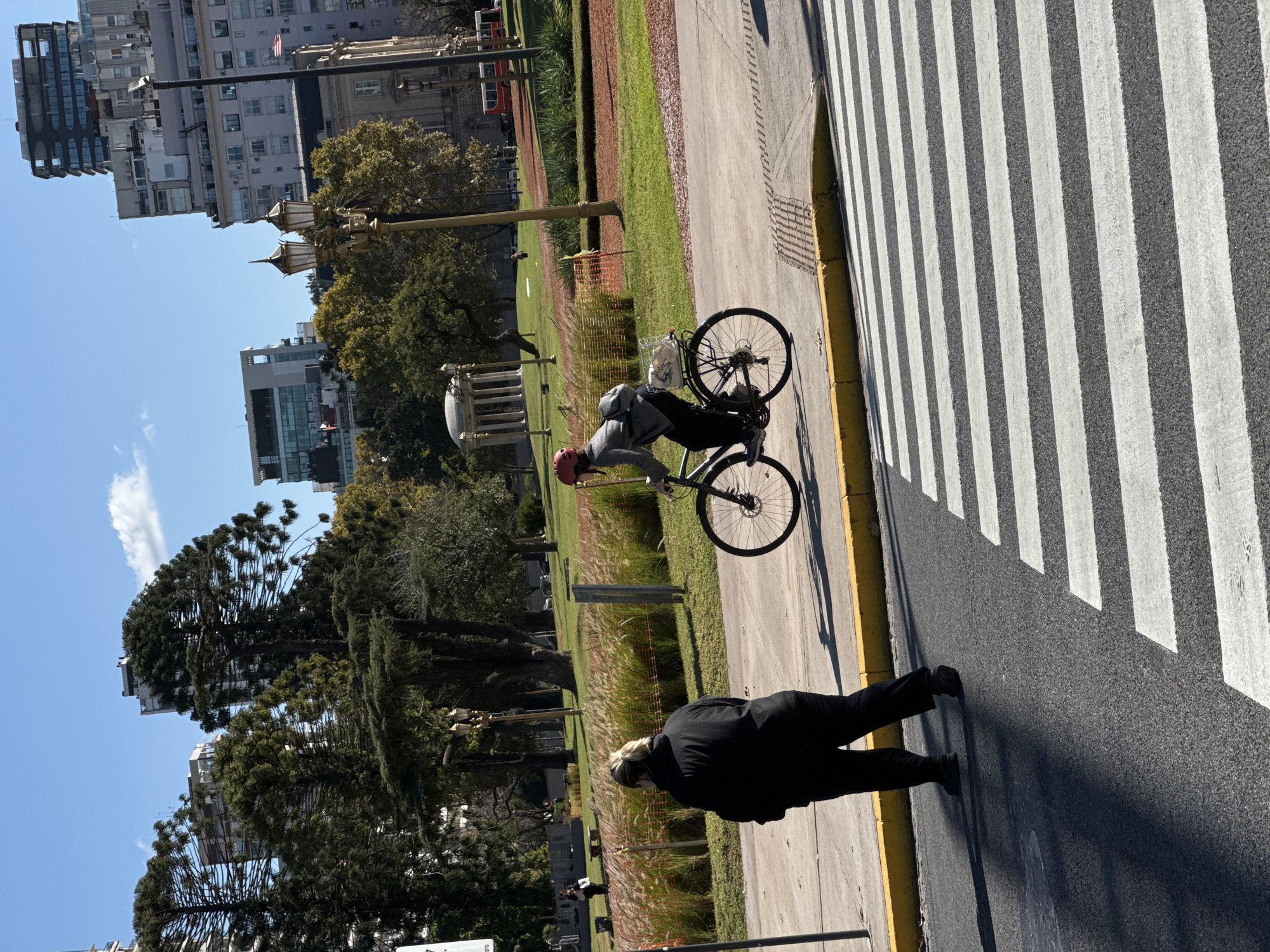 The image depicts a scene in Buenos Aires, Argentina. A person in a black outfit is walking along a sidewalk bordering a crosswalk. A cyclist wearing a grey sweater and a red helmet rides on the sidewalk next to a grassy area. In the background, trees and buildings are visible under a blue sky. The scene seems to be in a park or a green area within the city.
