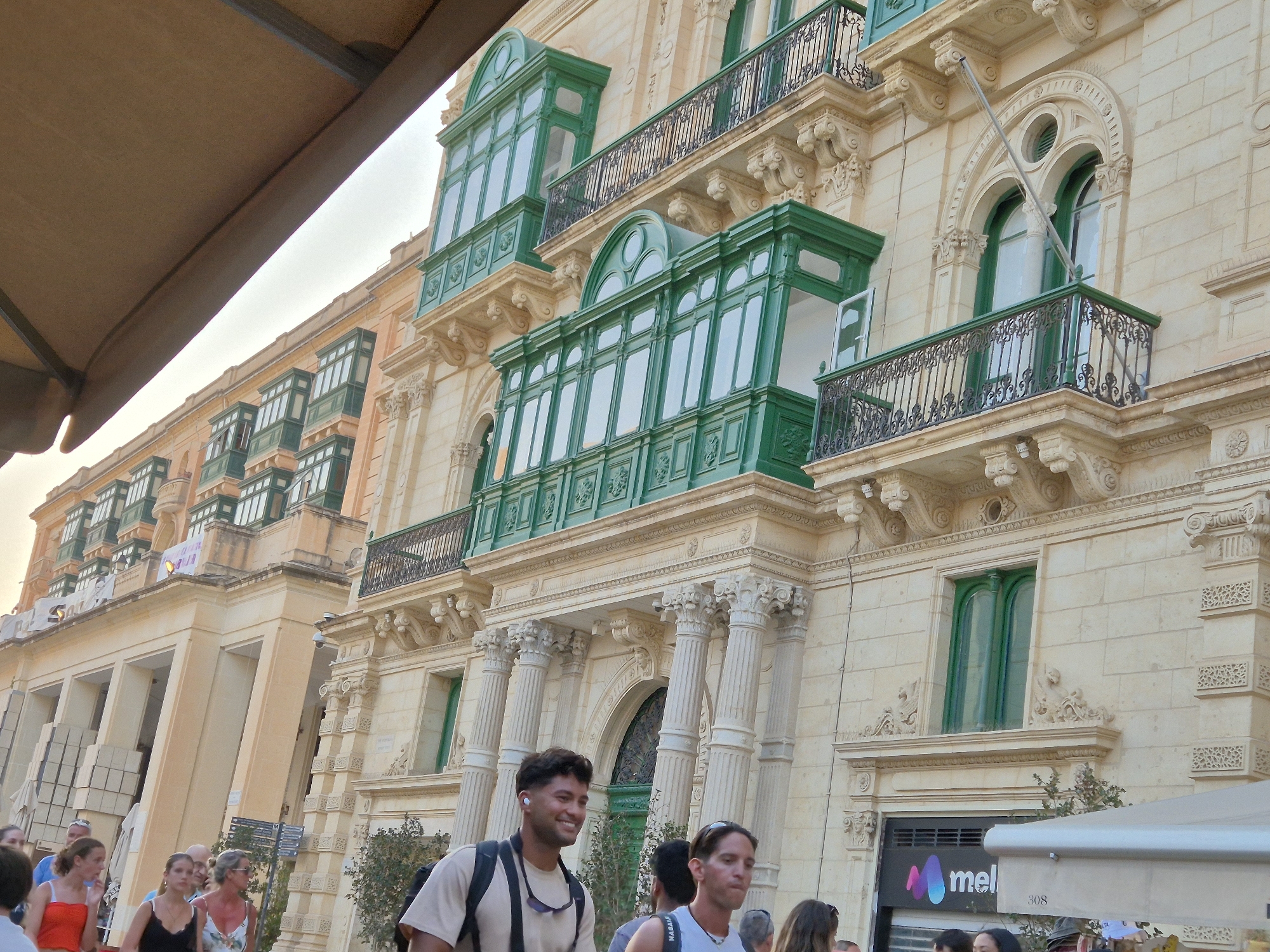 The image captures a street scene in Valletta, Malta, focusing on the distinctive architecture of buildings characterized by their ornate balconies. In the foreground, a man with dark curly hair and white earbuds smiles as he walks, wearing a light brown t-shirt and a backpack. To his right, another man with dark hair and a white t-shirt walks past, his gaze directed forward. Other individuals are partially visible in the background, suggesting a public space with pedestrian activity.
The architecture of the buildings is a prominent feature, with multi-story structures adorned with enclosed balconies, many painted in a vibrant green. These balconies, a common sight in Valletta, are a defining element of the cityscape. The buildings are constructed from a light-colored stone, common in Malta, and feature classical architectural details such as columns, arches, and decorative carvings. The sky appears overcast or hazy. A partially visible beige awning stretches across the top left of the frame, and a white tent-like structure is present on the right, indicating commercial activity or outdoor seating. A sign with the word "mel" and colorful abstract shapes is visible on a storefront. The overall impression is a lively urban environment showcasing Valletta's unique architectural heritage.