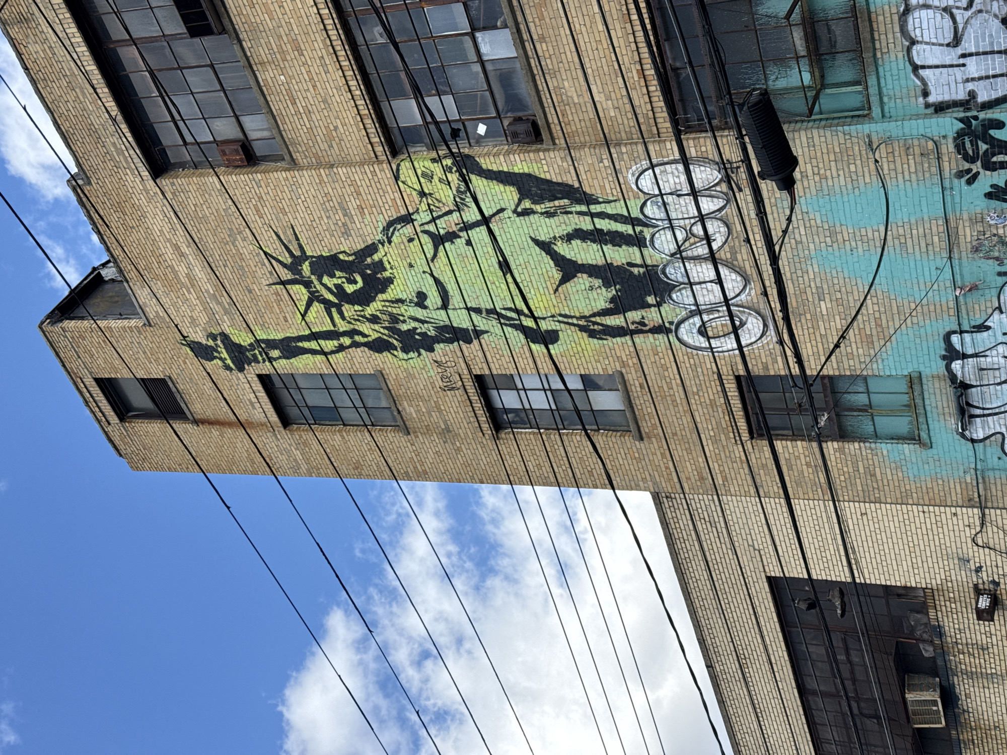 The image shows a low-angle view of a brick building in New York City. The building's exterior features several windows and extensive graffiti, including a depiction of the Statue of Liberty and other abstract designs. Overhead, a blue sky with scattered clouds is visible. Numerous power lines crisscross the view, adding to the urban scene.