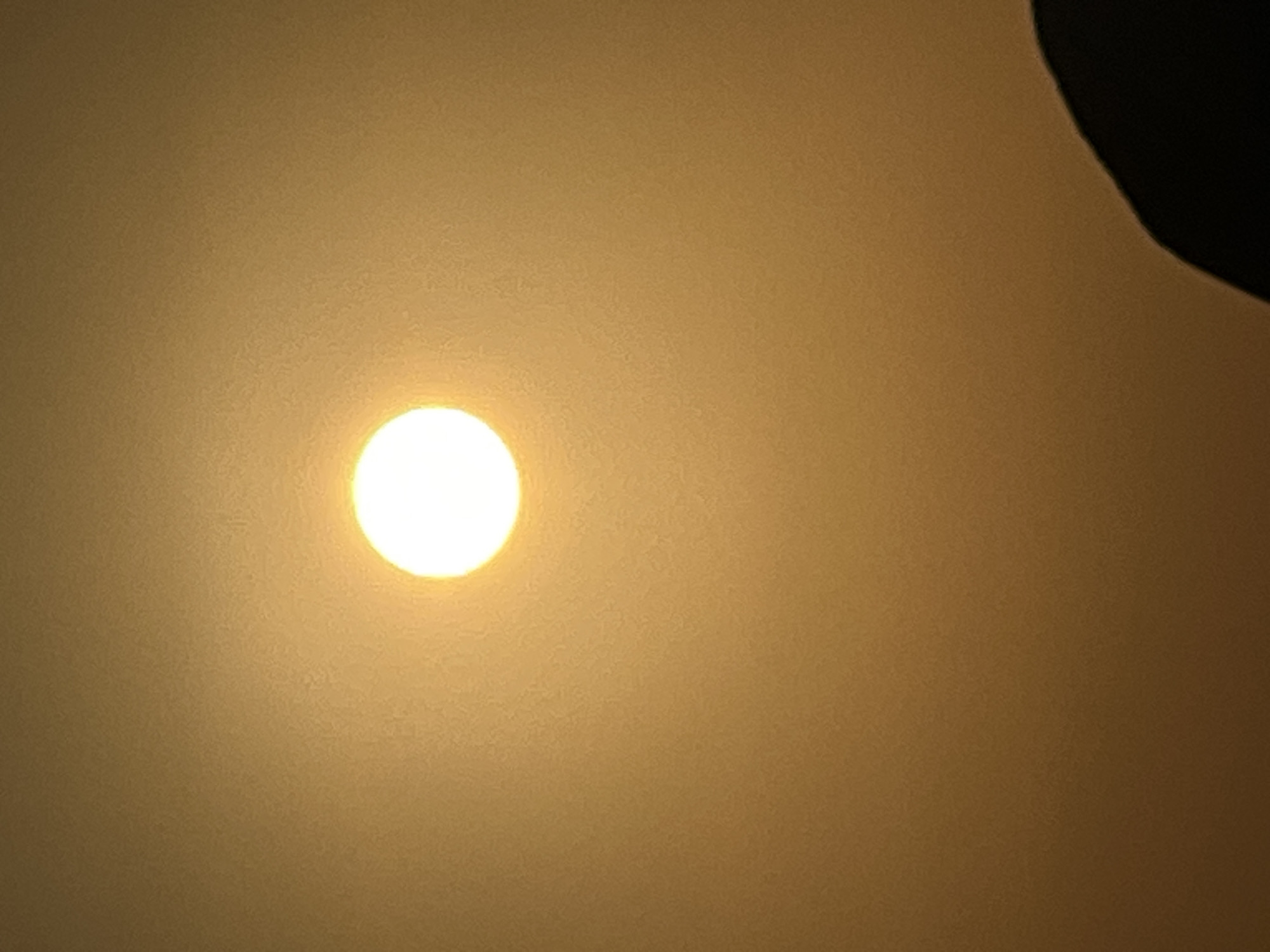 A bright, circular orb, possibly the sun, is centrally located in an otherwise undifferentiated, hazy, yellowish-orange sky. The lower right portion of the image is dominated by a dark, curved shape that appears to be the edge of a silhouette, potentially a person's head and shoulders, seen from behind. The scene is abstract and lacks specific urban context, making it impossible to determine the location within Kuta, Indonesia.