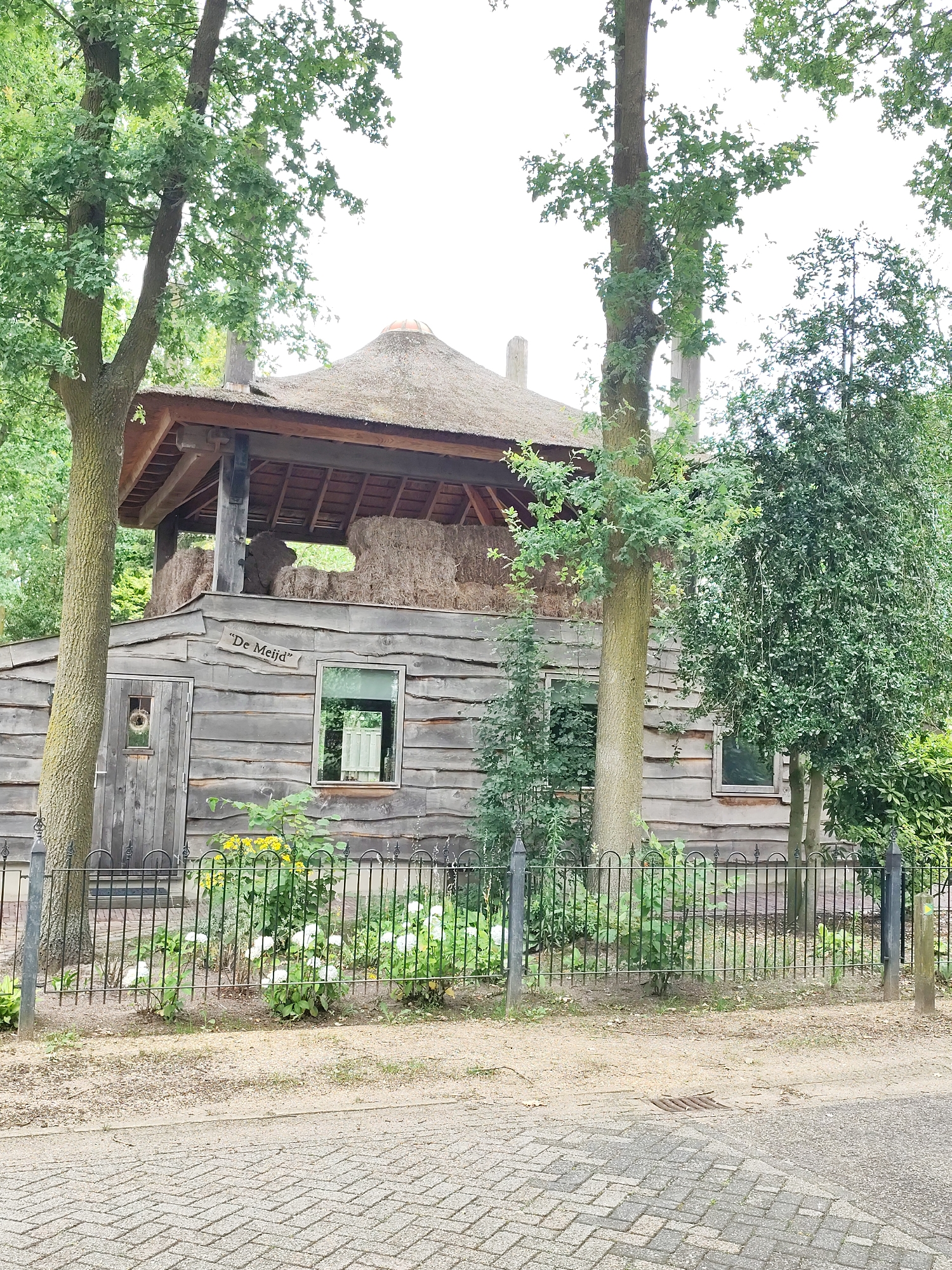 A wooden building with a thatched roof, appearing to be a barn or storage structure, is situated in Schaijk, Netherlands. The building has wooden plank siding and a covered upper level filled with bales of hay. A sign with the words "De Meijd" is affixed to the front of the building. A black metal fence encloses a garden area in front of the building, which features yellow and white flowers. Trees surround the structure, and a cobblestone path is visible in the foreground. There are no people or animals in the image.