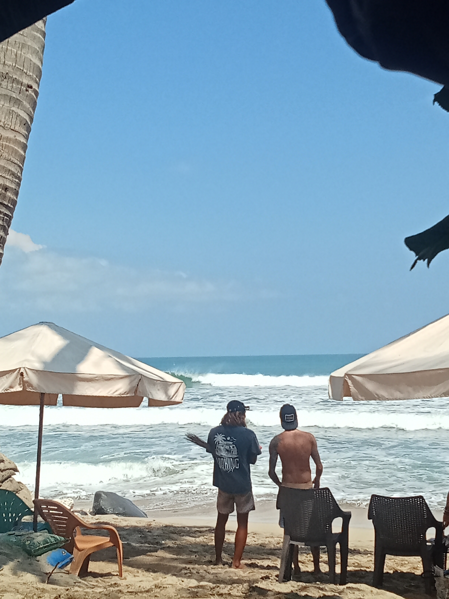 The image depicts a beach scene in Kuta, Indonesia, on a clear, sunny day. Two young men, likely surfers, are standing on the sand, facing the ocean. The man on the left wears a dark t-shirt with a graphic print and light-colored shorts, with a baseball cap on his head. The man on the right is shirtless, wearing light-colored shorts and a baseball cap, with his back to the camera. They appear to be observing the waves, which are moderate in size.
Two large beach umbrellas, one a light beige color and the other partially visible and also light in color, provide shade on the sandy beach. In the foreground, there are several plastic chairs, one brown and two dark grey, scattered on the sand. A brown plastic chair is positioned to the left of the two men, and two dark grey chairs are in front of the man on the right. Some rocks and foliage are also present on the beach. The ocean is a vibrant blue, with white-capped waves rolling towards the shore. A tall palm tree trunk is visible on the far left of the frame, and a dark silhouette of foliage hangs from the top right corner, suggesting the presence of trees or structures overhead. The overall atmosphere is one of a relaxed beach day.