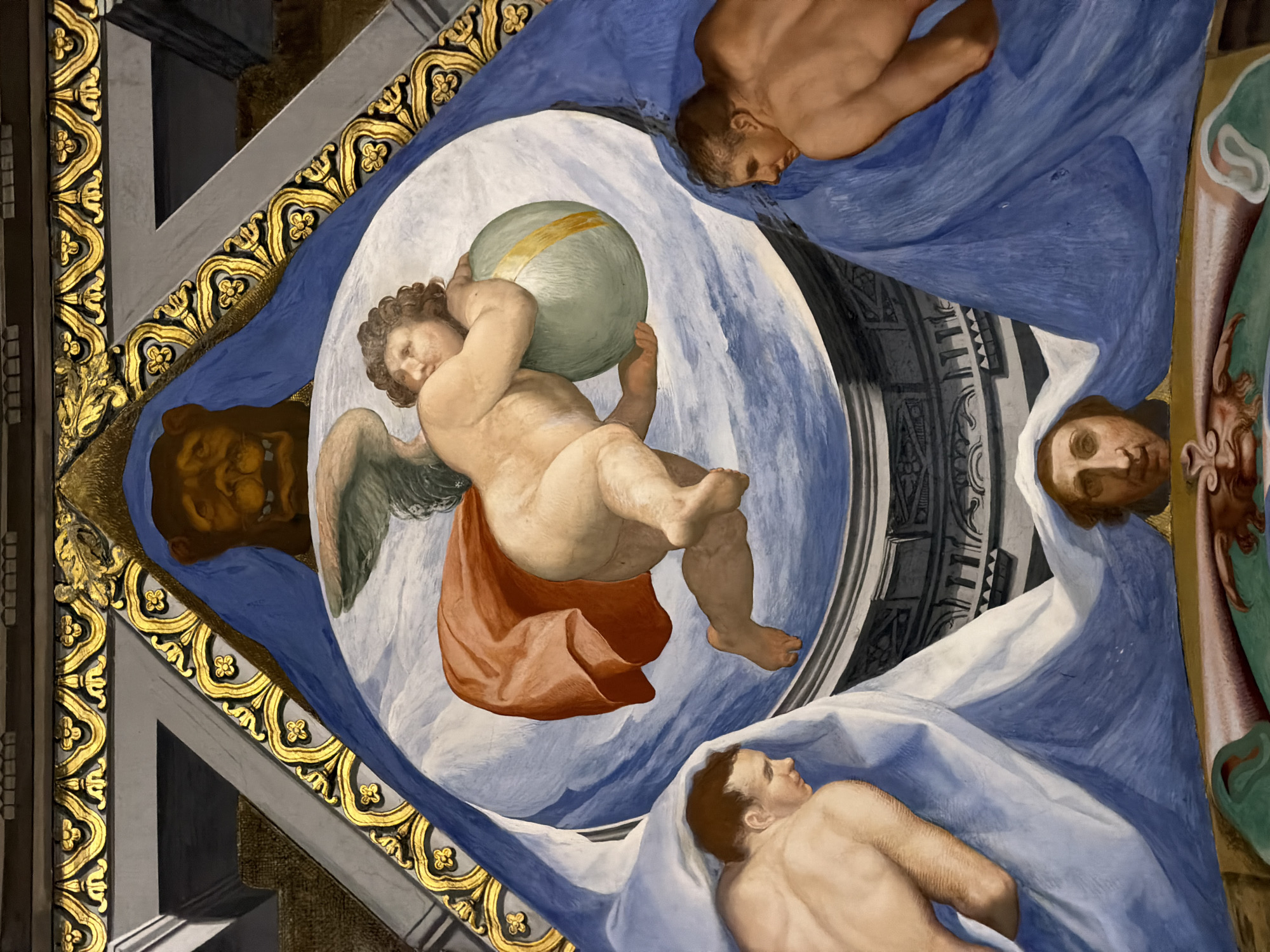 This is a detail from a fresco painting located in the Royal Monastery of San Lorenzo de El Escorial, Spain. The scene depicts a cherubic winged figure in the center, holding a light green globe with a golden band. The figure is rendered with soft, fleshy skin, and is draped in a vibrant orange cloth that billows behind it.
To the left and right of the central figure are the upper torsos of two male figures, draped in blue fabric. Their skin tones are rendered with a warm, bronzed hue. Their heads are turned towards the central cherub.
At the bottom of the composition, a human face emerges from beneath blue drapery, looking upwards. Above the central cherub, a lion's head is depicted within a brown, stylized frame. The entire composition is framed by ornate gold and grey architectural elements, suggesting it is part of a larger ceiling or wall decoration. The overall style is classical and dramatic, typical of Renaissance or Baroque art.