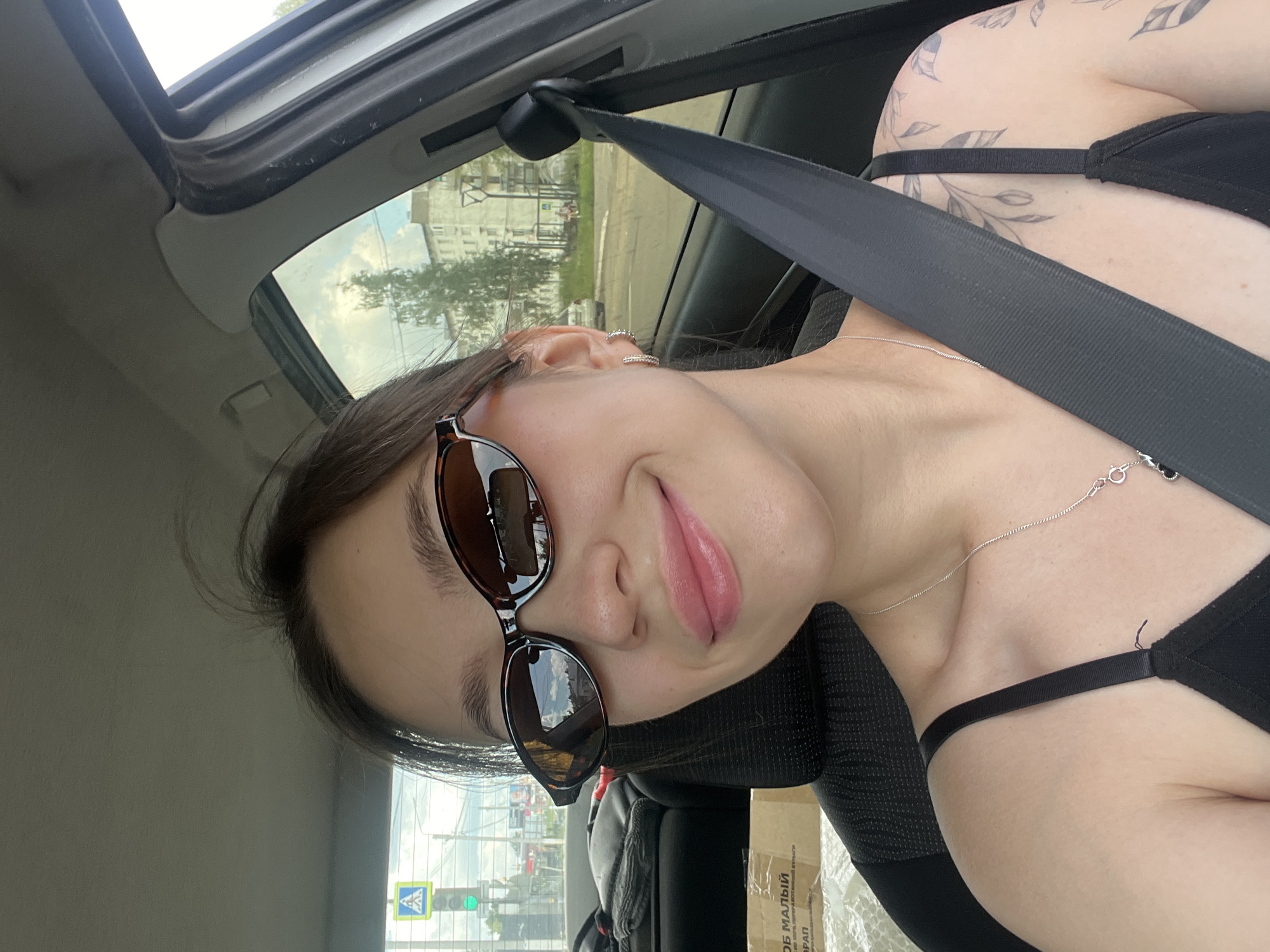 A young woman with dark hair and sunglasses is taking a selfie inside a car. She is wearing a black tank top and a silver necklace. A seatbelt is visible across her body. The background shows a city street with buildings and trees, suggesting an urban or suburban environment. A traffic light is visible through the windshield. The image appears to be taken during the daytime, with good lighting. The context suggests the woman is a resident of Chaikovsky, Russia.