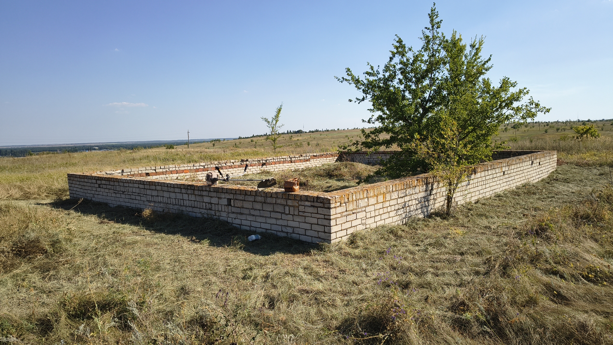 This image depicts the partially constructed foundation of a building made of light-colored bricks, located in a rural, grassy area under a clear blue sky. The foundation is rectangular in shape, with a mature tree with green foliage growing to the right of it, casting a shadow on the brickwork. Inside the foundation, there is a pile of hay or dried grass. Various construction tools and materials are visible within the foundation's perimeter, including what appears to be a trimmer, a shovel, and a terracotta-colored object. In the background, the landscape is characterized by rolling hills covered in dry grass, with some scattered trees and a distant line of trees on the horizon. A utility pole is visible in the far distance to the right. The scene suggests an unfinished construction project in an open, undeveloped area.