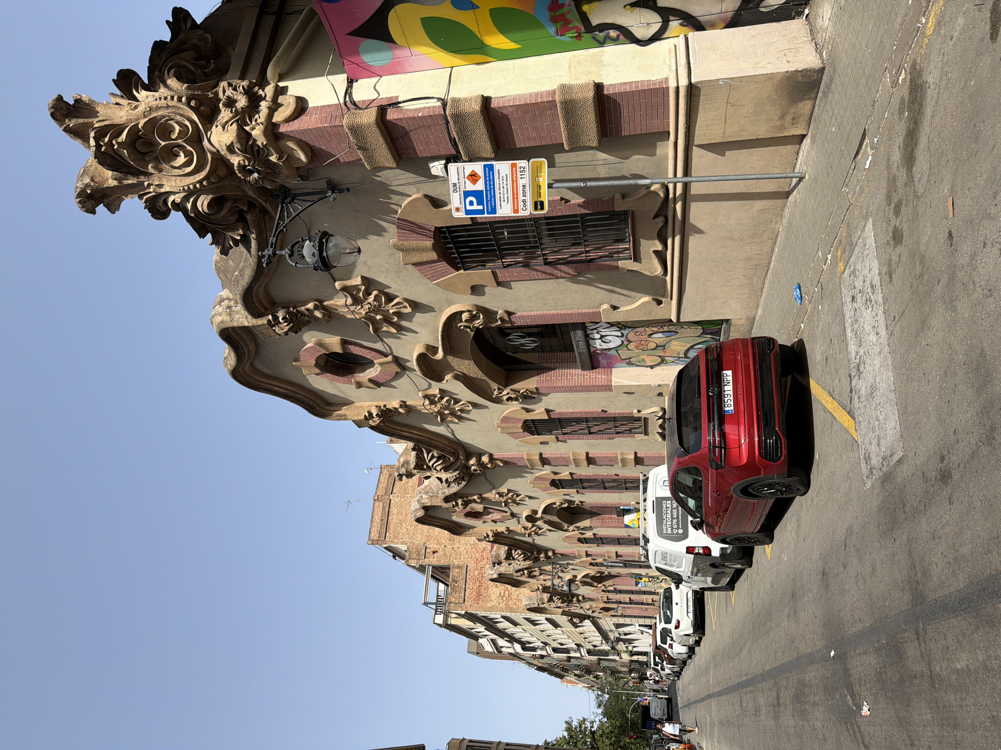 The image depicts a street scene in Barcelona, Spain, with a focus on the exterior of a building featuring ornate architectural details. The building's facade is painted a light tan color and adorned with intricate stucco carvings, including decorative motifs and what appear to be stylized faces. Red brickwork is visible around the windows and as accent elements on the facade. Several windows are covered with black metal bars. Graffiti is present on the lower portions of the building and adjacent walls, featuring colorful abstract designs.
Parked along the street are several cars and a white van. The foreground shows a red Volkswagen SUV. A metal pole with a parking sign is positioned on the sidewalk. The sign has text in Spanish and indicates parking regulations. The street itself is paved and has yellow painted lines indicating parking areas. The sidewalk is made of interlocking paving stones. The sky is clear and blue, suggesting a sunny day. In the background, other buildings of varying heights and architectural styles are visible, some with exposed brick. A pedestrian is walking on the sidewalk in the distance.