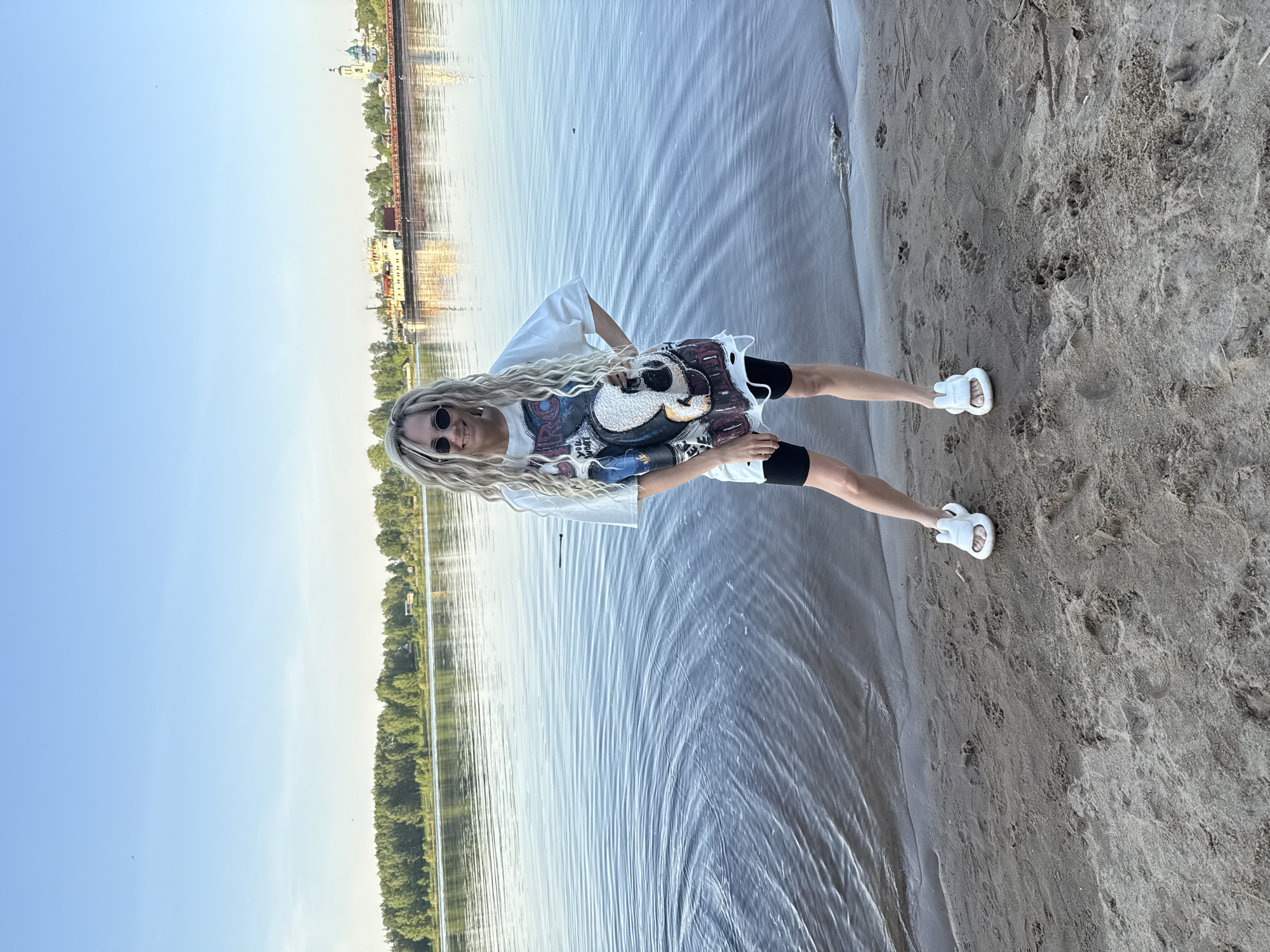 A young woman with blonde dreadlocks and sunglasses stands on a sandy beach, partially submerged in water. She wears a white t-shirt with a large graphic design, black shorts, and white sandals. Behind her, the image shows a wide expanse of water, possibly a river or lake. On the opposite bank, there's a cityscape visible, featuring buildings, trees, and potentially some industrial structures. Given that the image is taken in Rusanovo, Russia, the cityscape would likely be part of that city. The sky is mostly clear with a bluish hue.