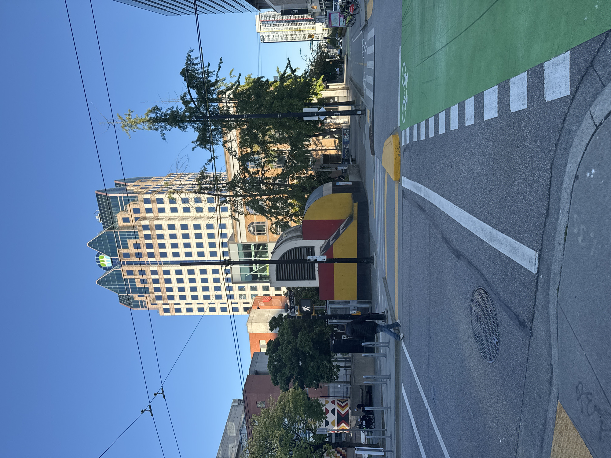 The image shows a street scene in Vancouver, Canada, with a tall, light-colored building with a distinctive faceted glass roof as the dominant structure. In the foreground, a pedestrian crosses a street marked with crosswalks and a green-painted bike lane. A person walks from left to right across the intersection. The street is wet in places, suggesting recent rain or dew. Overhead wires crisscross the sky, common in older city districts. Other buildings of varying heights and architectural styles are visible, along with trees and street furniture. A red and yellow structure, possibly a public art installation or part of a building's facade, is located near the pedestrian crossing. The sky is clear and blue, indicating a sunny day. The overall impression is of a busy urban environment with elements of modern and older architecture.