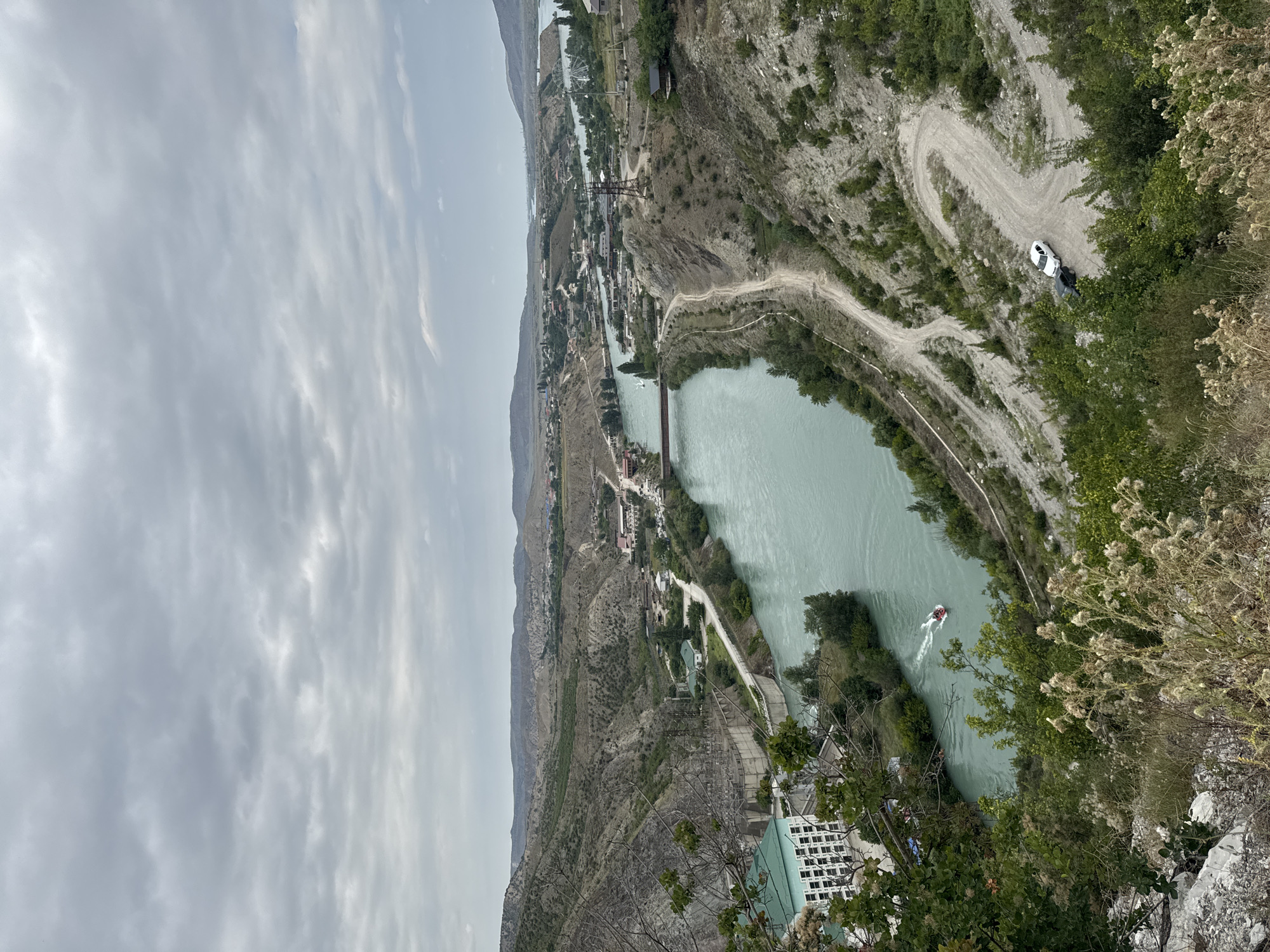 The image captures a broad, elevated view of a river valley with a body of water running through it. The river is a bright, turquoise color and appears to be wide. Along the banks of the river, there are various structures, including buildings with green roofs and a prominent dam or hydroelectric power station on the left side. A road winds along the right bank of the river, and two cars are parked on this road. A small red and white boat is navigating the river, leaving a white wake. The surrounding landscape is characterized by rolling hills and mountains with sparse vegetation, suggesting a dry or semi-arid climate. The sky is overcast with a layer of gray clouds. The location is identified as Novo-Zubutli, Russia.