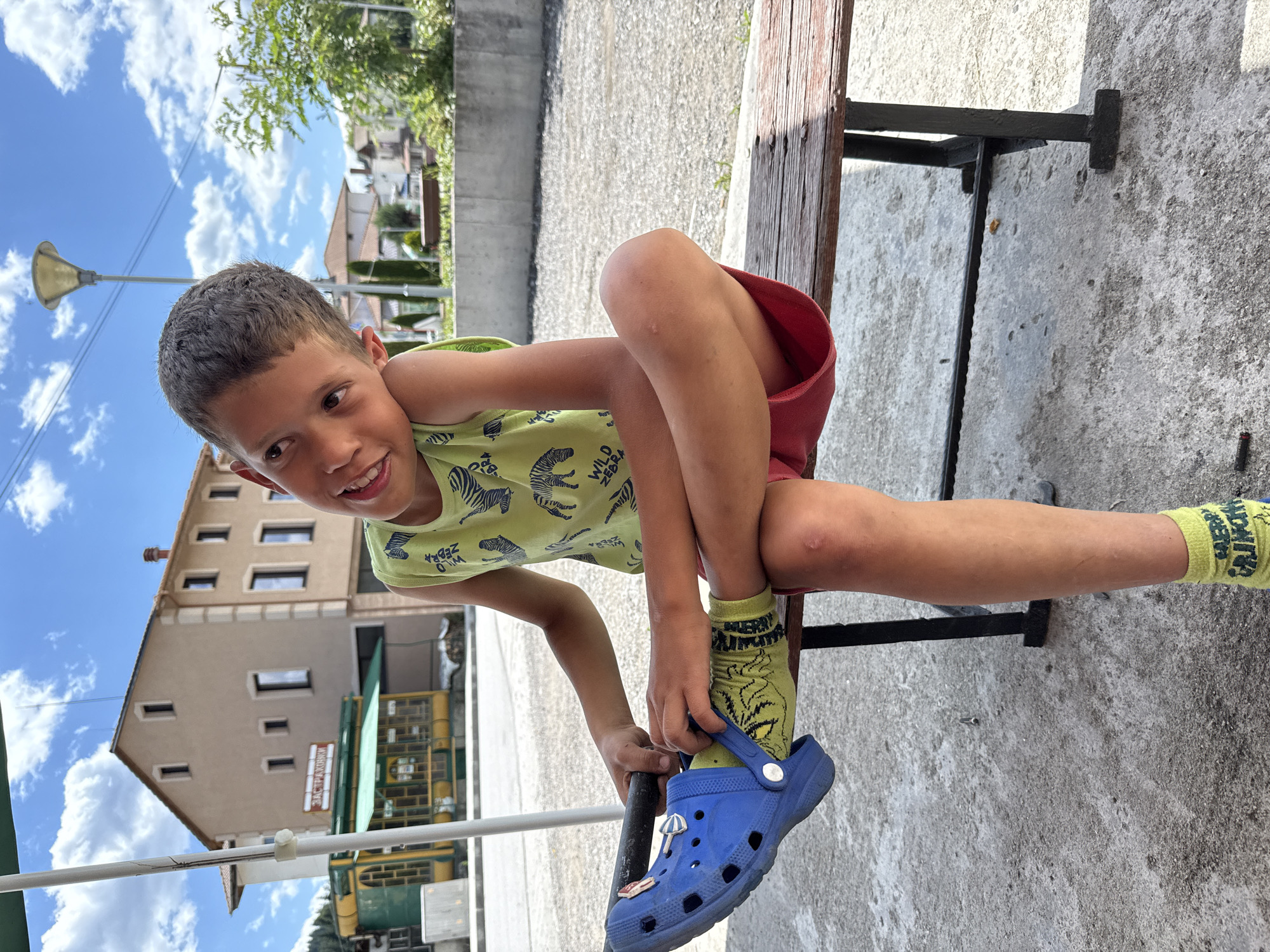 A young boy with short, dark hair is sitting on a wooden bench, looking at the camera with a wide smile. He is wearing a light green t-shirt with a zebra print and red shorts. His right leg is bent, and he is in the process of putting on a blue Croc shoe with a small beach-themed charm attached. He is also wearing yellow socks with a black pattern. The scene appears to be outdoors, possibly in a park or a public area, with a concrete ground and a building in the background. The sky is blue with white clouds, suggesting it is daytime and the weather is pleasant. A streetlamp is visible on the left side of the image. The building in the background has multiple windows and a sign with Cyrillic text that reads "ЗАСТРАХОВКИ" (which translates to "INSURANCE"). The overall mood of the image is cheerful and candid.