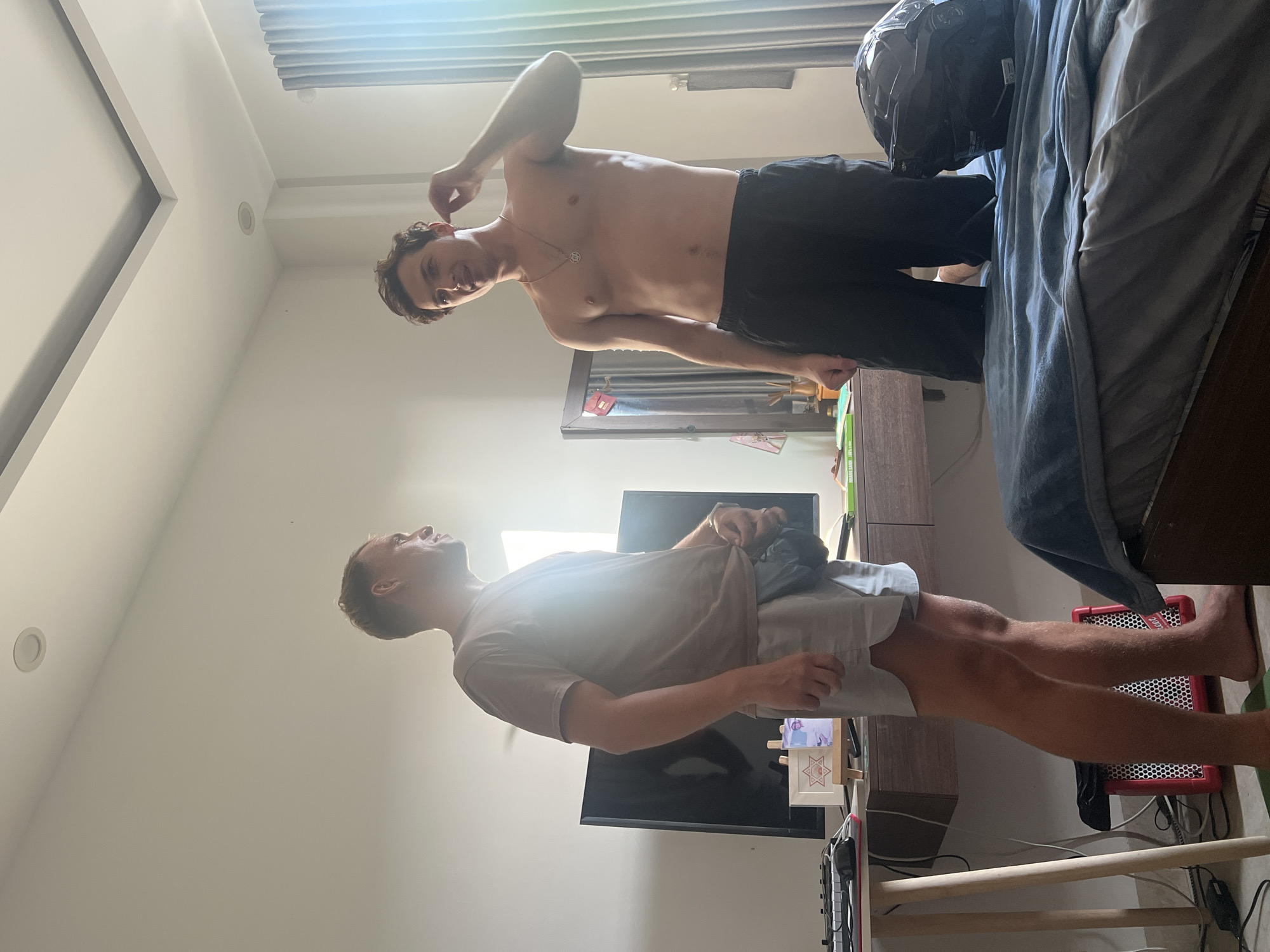 Two young adult males are in a bedroom in Đà Nẵng, Vietnam. The room is brightly lit by natural light coming from the right side of the frame. The walls are white, and the ceiling has recessed lighting.
On the left, a male with short, light-brown hair is standing and facing the right. He is wearing a light-colored t-shirt and light-colored shorts. He has a black fanny pack around his waist. He is looking towards the other male.
On the right, a shirtless male with dark, curly hair is standing and facing the left. He is wearing dark shorts. He has a necklace with a star pendant. He is gesturing with his right hand towards his head.
In the background, there is a black flat-screen television on a wooden desk. The desk also has a keyboard, a small framed picture, and some books. To the right of the desk, there is a mirror. A motorcycle helmet sits on a bed covered with a blue blanket. Curtains are visible on the far right. The overall impression is a casual, domestic setting.