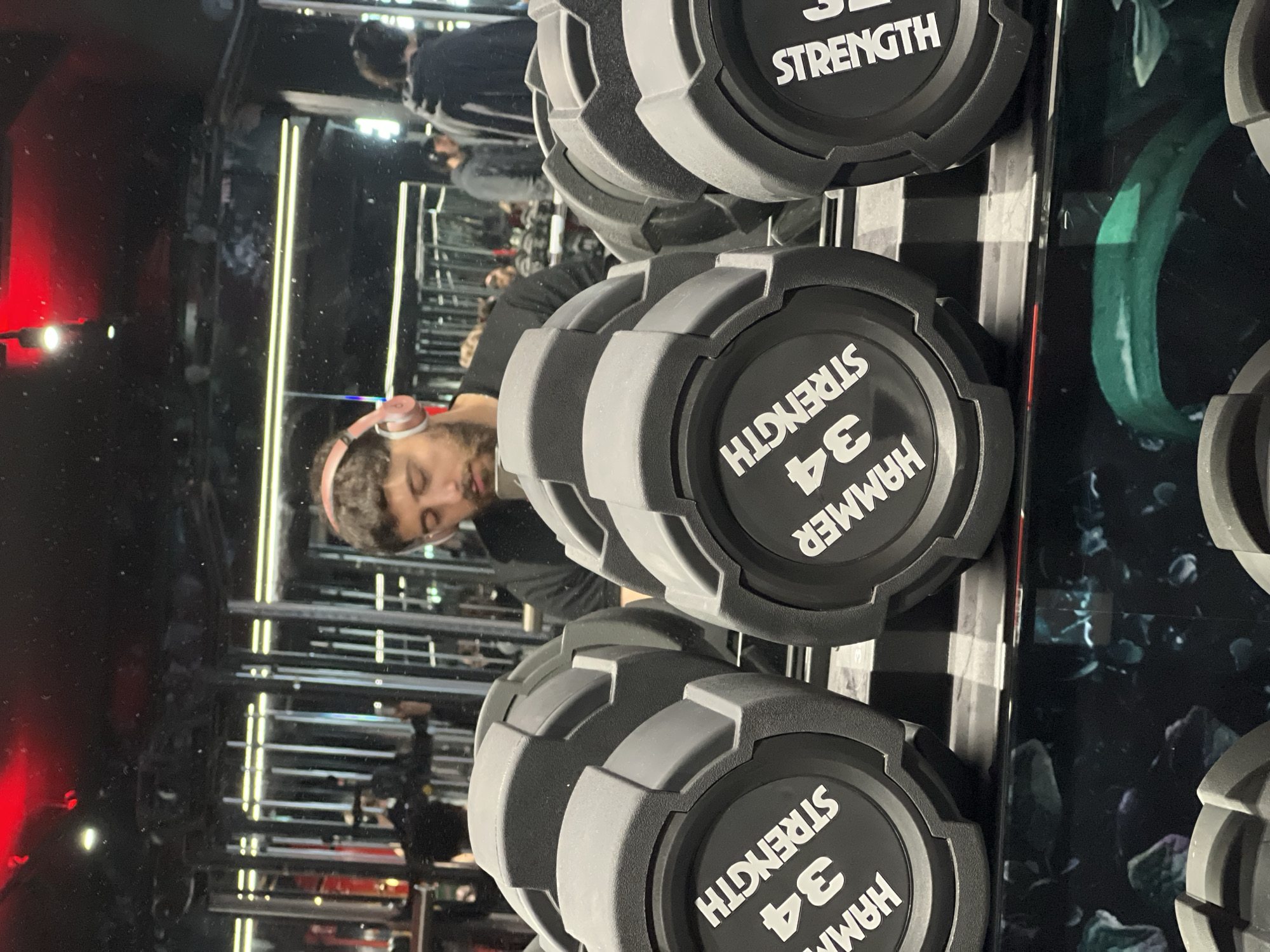 The image shows a rack of dumbbells in a gym. The dumbbells are labeled "HAMMER STRENGTH 34". In the background, a person with headphones is reflected in a mirror. The gym appears to have mirrored walls and modern lighting. While the location cannot be definitively determined to be Strasbourg, France based on visual evidence alone, the image depicts a gym environment.
