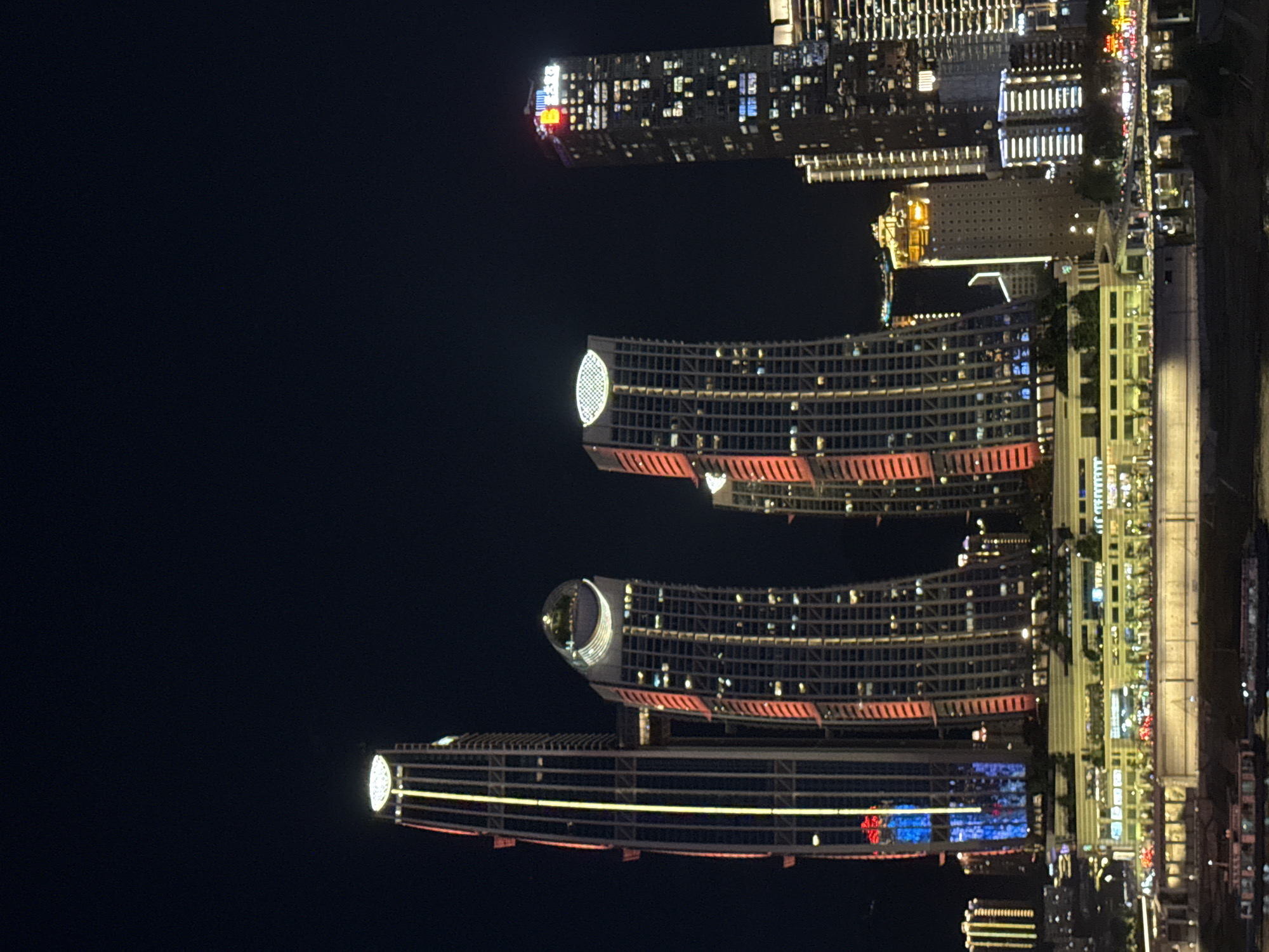 The image shows a nighttime view of a city skyline, specifically 江北城街道, China, with numerous illuminated skyscrapers and a waterfront area. The buildings are of varying heights and architectural styles, with many featuring colorful LED lighting that outlines their structures and windows. A prominent feature is a cluster of tall, modern buildings in the foreground, some with distinctive curved or angled designs. One skyscraper on the left is lit with vertical yellow and red lights, while adjacent buildings have red and white lights. A curved building in the center has a circular, illuminated structure at its apex. To the right, another tall skyscraper displays a red and yellow logo at its top, and its windows are lit in white and blue. In the mid-ground, a long, low-rise building complex runs along the waterfront, also brightly lit with numerous windows and some green landscaping. Several boats are visible on the water in the foreground, and a bridge with traffic lights is also present. The sky is dark, indicating the scene is taking place after sunset. No people are discernibly visible in the image.