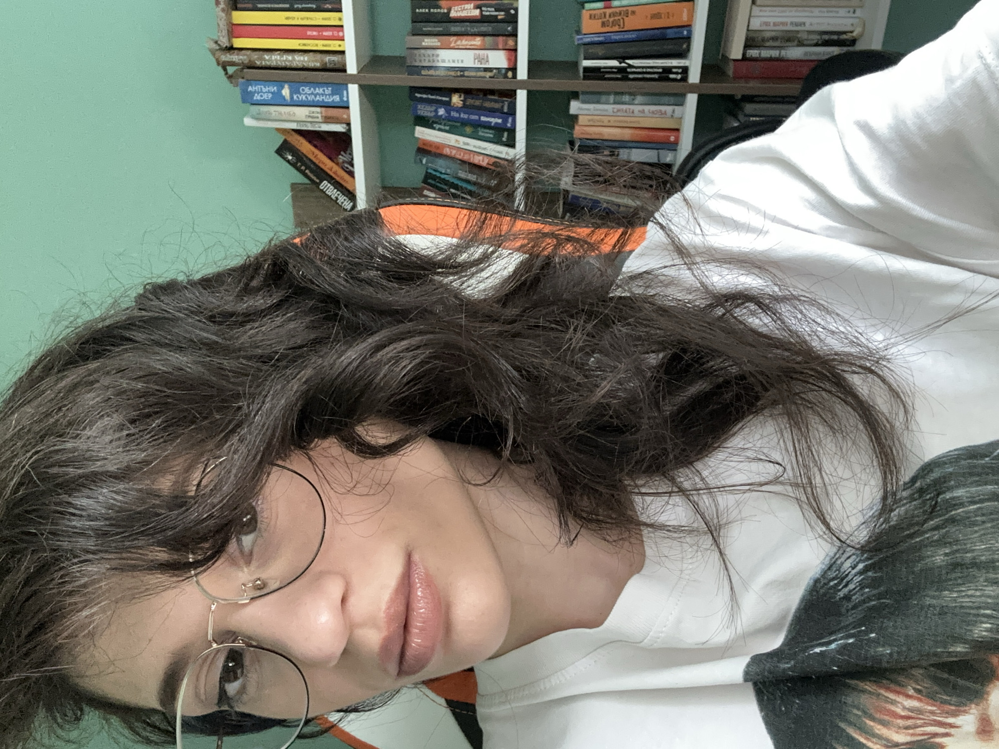 The media file is a close-up, head-and-shoulders shot of a young person with long, dark, wavy hair. They are wearing large, round, thin-rimmed glasses and a white hooded sweatshirt with a graphic print of a person's face on the front. The person's lips are slightly parted and appear to be glossed. The background features a shelf filled with books, some of which have visible text in Cyrillic script. The wall behind the bookshelf is painted a mint green. The lighting suggests it is daytime, and the overall mood is relaxed and informal, possibly a selfie taken at home. The presence of Cyrillic text on the books, combined with the provided location context of Sofia, Bulgaria, indicates the setting is likely in Bulgaria.