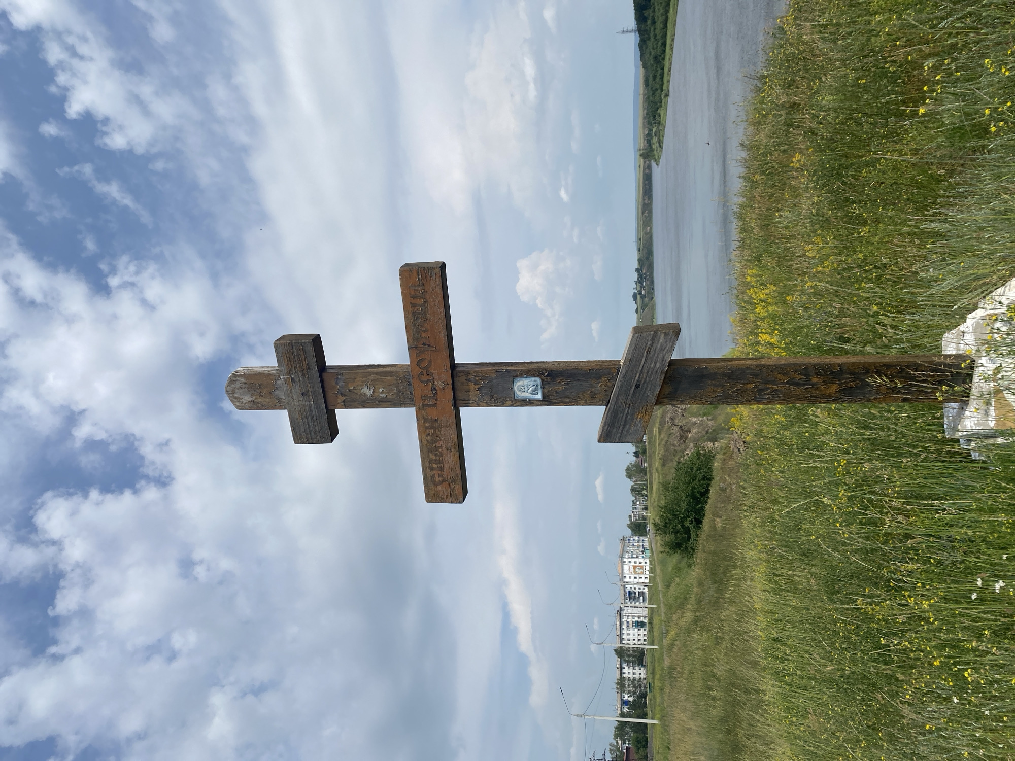 A wooden cross stands prominently in the foreground of the image, its weathered wood and simple construction suggesting a religious or memorial purpose. The cross is positioned on a grassy hilltop overlooking a wide river that curves into the distance. Behind the cross, a cluster of apartment buildings and some utility poles are visible, indicating a nearby urban or suburban area. The sky is partially cloudy, with patches of blue sky interspersed with white cumulus clouds. The surrounding landscape is characterized by rolling hills covered in green grass and wildflowers. The overall scene appears to be set in a rural or semi-rural location, possibly on the outskirts of a town. The image is taken from a low angle, emphasizing the height of the cross against the sky and the expanse of the landscape. The presence of the buildings suggests that this location is within or near the city of Novomaltinsk, Russia.