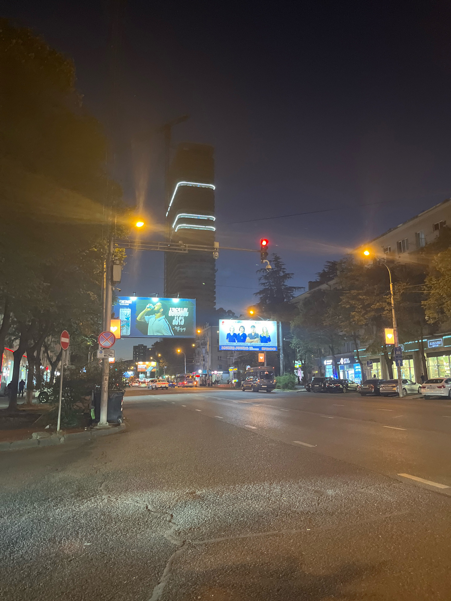The image depicts a nighttime street scene in Tbilisi, Georgia, likely on Rustaveli Avenue, based on the presence of prominent billboards and a distinctive modern skyscraper. The perspective is from the middle of a paved road, looking forward. The sky is dark, indicating it's late evening or night.
On the left side of the road, there are trees and building facades, with some of them appearing to be shops or businesses with illuminated interiors. Traffic lights are visible, with one showing a red light. There are several billboards on the left, one featuring a man drinking from a bottle and text in Georgian script, and another with three individuals.
Further down the road, a tall, modern skyscraper with illuminated white bands is a prominent landmark. The street is lined with trees and various buildings. Several cars are present on the road, some parked and others in motion. The road surface appears to be asphalt and shows signs of wear.
On the right side of the road, more buildings are visible, including one with multiple windows and balconies. Traffic lights are also present on this side. The overall impression is a busy urban environment at night, with illuminated advertisements and traffic contributing to the scene.