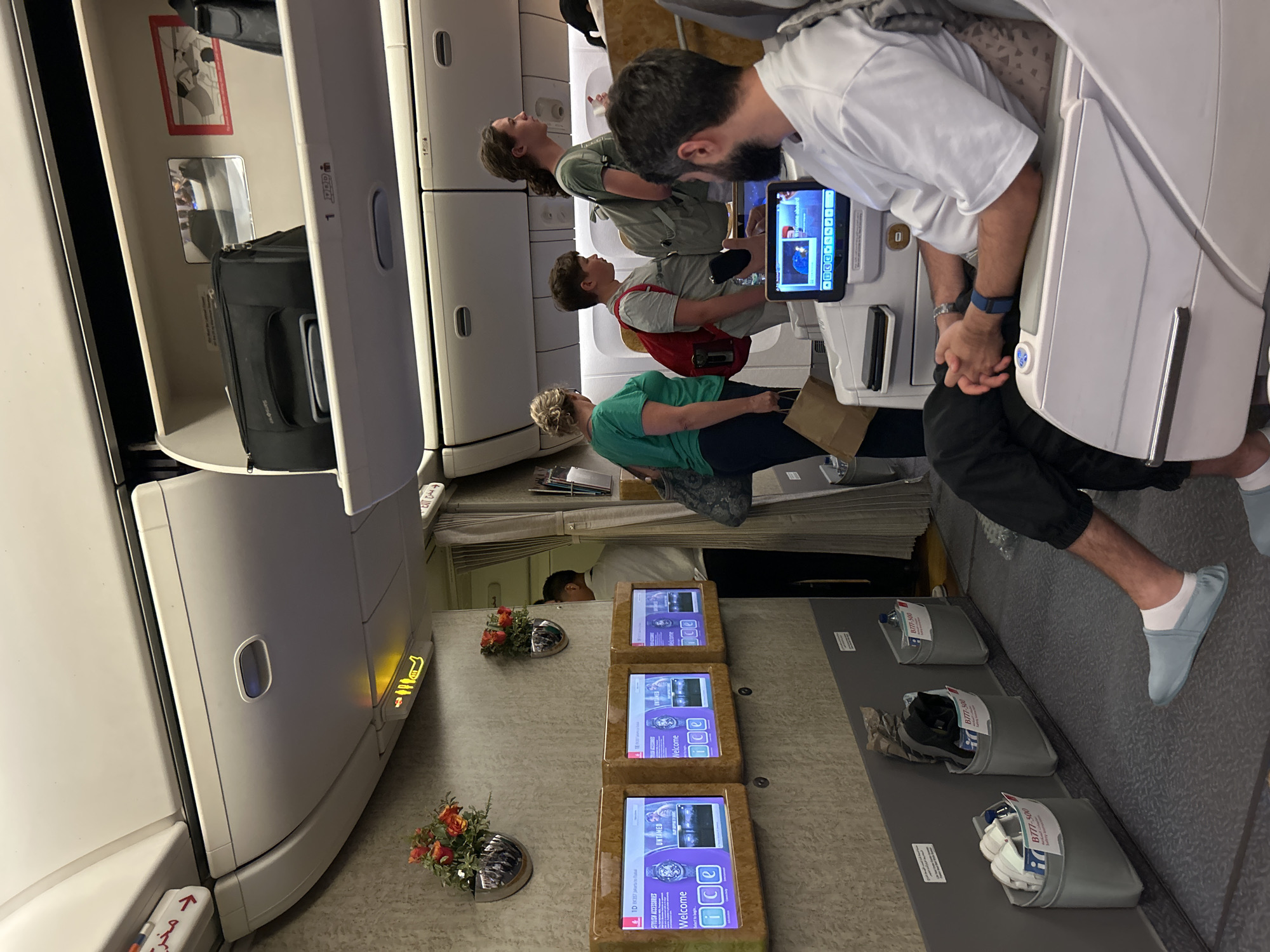 The image depicts the interior of an airplane cabin, possibly first or business class, given the spacious seating and amenities. In the foreground, a man is seated in a large, reclined chair, facing a personal entertainment screen. He is wearing a white t-shirt, black pants, and light blue slippers. His hands are clasped in his lap, and he is wearing a watch and a blue fitness tracker. To his left, a section of the cabin features three wall-mounted screens displaying welcome messages and advertisements. Further down the aisle, a flight attendant in a white uniform stands near an open doorway. Several passengers are visible standing and walking in the aisle, including a woman in a green shirt, a boy with a red backpack, and another person in a grey t-shirt with a backpack. Overhead compartments are open, containing luggage. The cabin's decor features beige textured walls and overhead panels. The overall environment suggests a commercial flight in progress or about to depart. There is no indication that this scene is located in Tangerang, Indonesia.