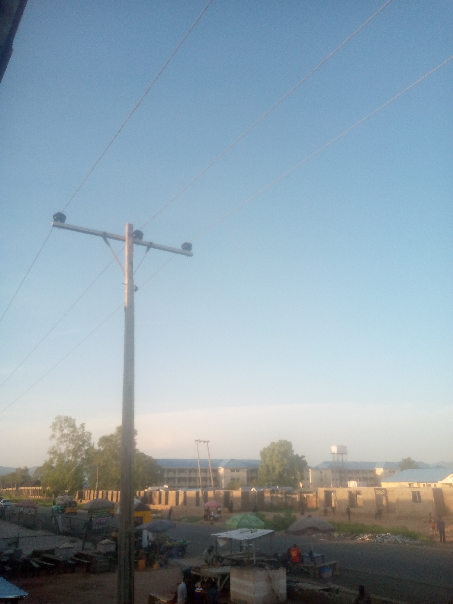 The image presents a daytime scene from the city of Jalingo, Nigeria, characterized by a pale blue, clear sky. A wooden utility pole with a horizontal cross-arm supporting three insulators and multiple power lines dominates the left-center of the frame, with lines extending diagonally towards the upper right.
The lower half of the image features a bustling street or roadside environment. An unpaved shoulder borders a dark, possibly asphalt, road. Numerous makeshift stalls, some covered by various colored umbrellas (including green and black), line the thoroughfare. Multiple individuals are scattered throughout the scene, engaged in unidentifiable activities around these commercial structures, suggesting a market or busy roadside.
In the midground, several structures are visible. On the right, buildings appear to be under construction or incomplete, featuring exposed block walls. Further back, a series of multi-story buildings with light-colored facades and dark roofs extend horizontally across the frame. To the far right, a tall water tower with a prominent tank stands against the sky. Various trees with green foliage are interspersed within the urban landscape. Distant hills or mountains are faintly discernible on the horizon in the background. The lighting suggests either early morning or late afternoon.