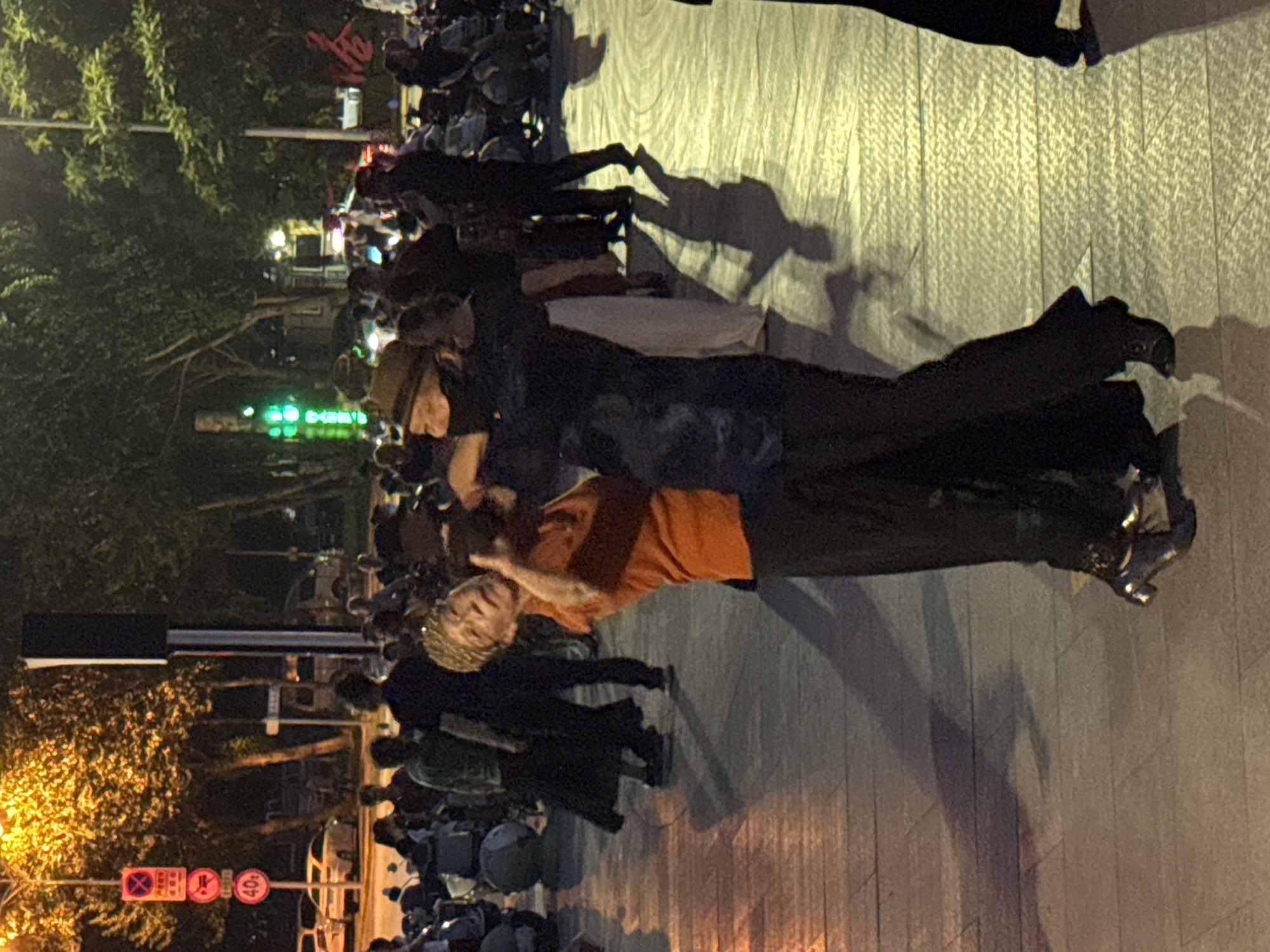 The image depicts a nighttime street scene in Jinzhou, China, possibly near a pedestrian area. In the foreground, a man and a woman are dancing. The woman, on the left, wears an orange shirt and black pants. The man, on the right, wears a dark jacket and a hat. They are in motion, with the woman leaning back. The ground is paved with textured tiles, reflecting the ambient light from streetlights and possibly nearby buildings. In the background, a crowd of people are visible, some seated and others standing, suggesting a public gathering or event. Trees line the street, and traffic lights and signage are present, including a sign with "40" indicating a speed limit. The overall atmosphere appears lively and social.