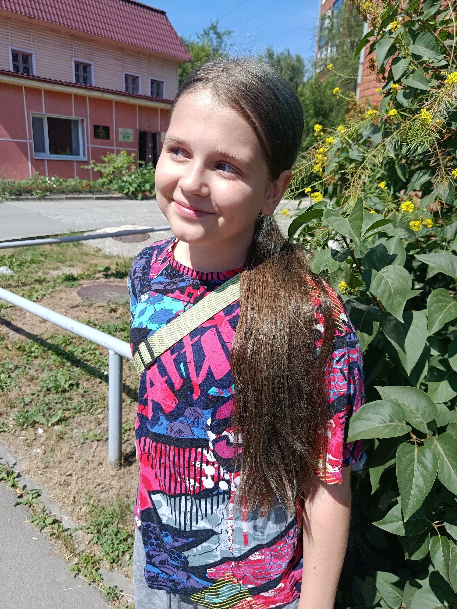 A young girl with brown hair in a ponytail, wearing a brightly colored t-shirt and grey pants, stands outdoors. She is smiling and looking to her left. The background shows a building with a red roof, green bushes with yellow flowers, and a sidewalk. This image was taken in Biysk, Russia.