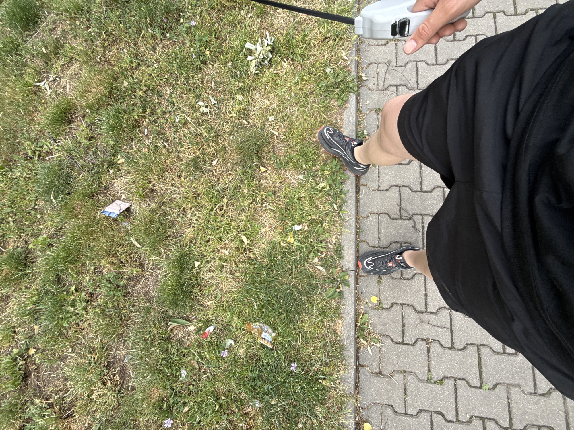 A first-person perspective photograph shows a person standing on a gray brick sidewalk adjacent to a grassy area containing trash. The person is wearing black shorts and black running shoes with red accents. A gray retractable dog leash is held in the person's right hand. The grassy area contains litter, including wrappers and other debris. The location is Stara Zagora, Bulgaria.