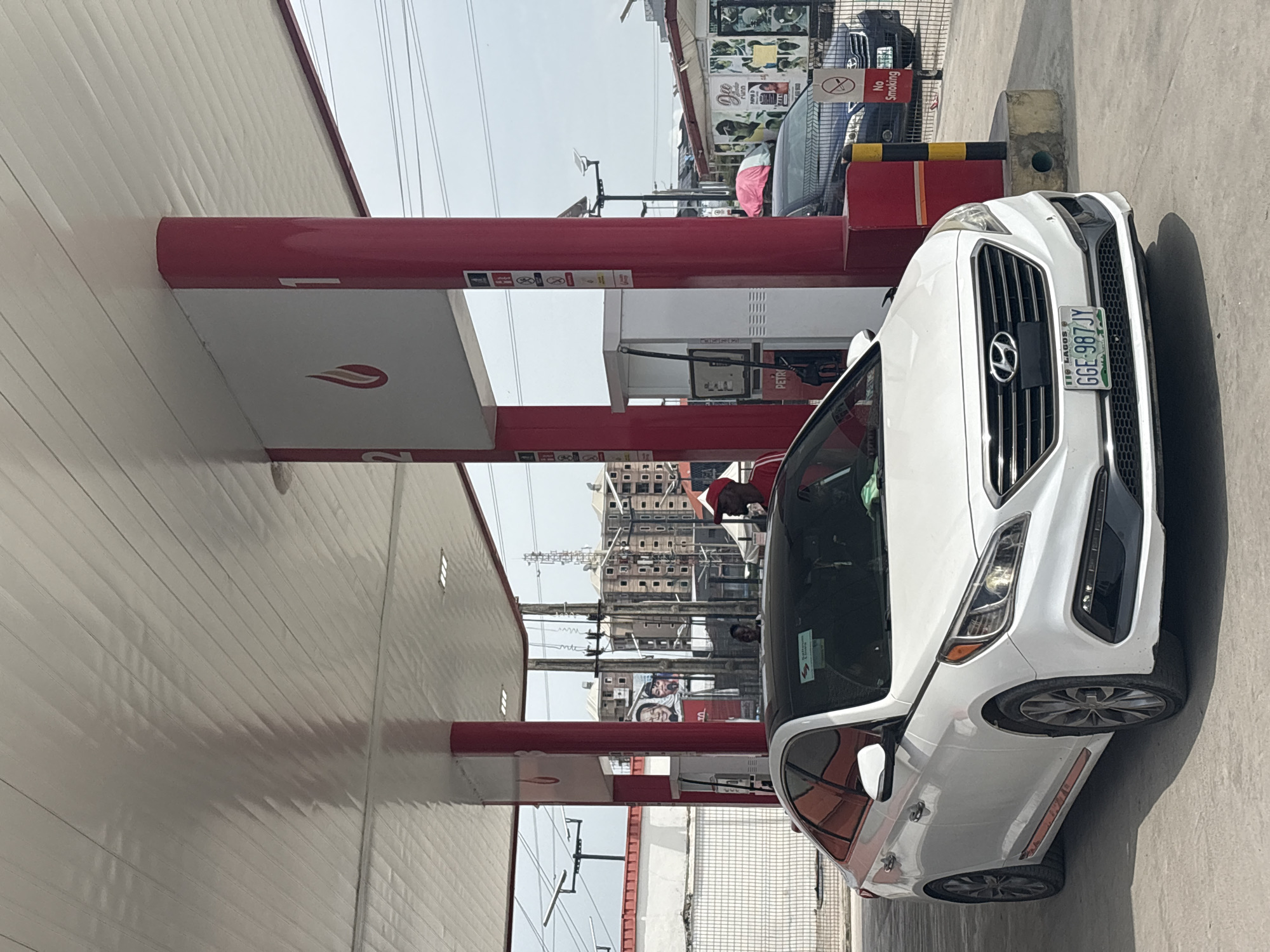 The image depicts a white Hyundai Sonata at a petrol station, likely in Igboefon, Nigeria, as indicated by the Lagos license plate "GGE-987JY". The car is positioned to the right of a red and white gas pump. In the background, buildings and power lines are visible, and a "No Smoking" sign is near a black vehicle parked behind the Sonata. A person wearing a red cap can be seen inside the car. A billboard is in the background.