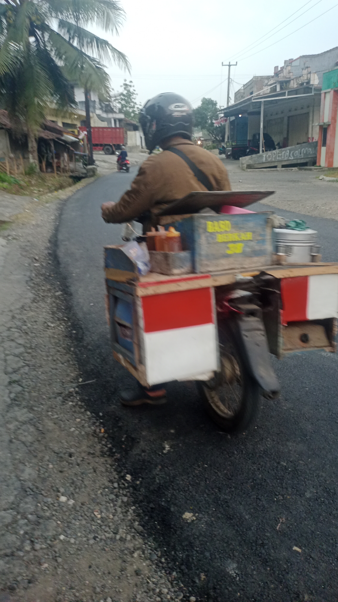 A motorcyclist wearing a brown jacket and a black helmet is riding on a paved road. The motorcycle is equipped with a cart carrying supplies, including bottles of sauce and what appears to be food containers. The cart has red and white sections, resembling the Indonesian flag. In the background, there are buildings, a red truck, and palm trees. The scene suggests a typical street view in Cogreg, Indonesia.