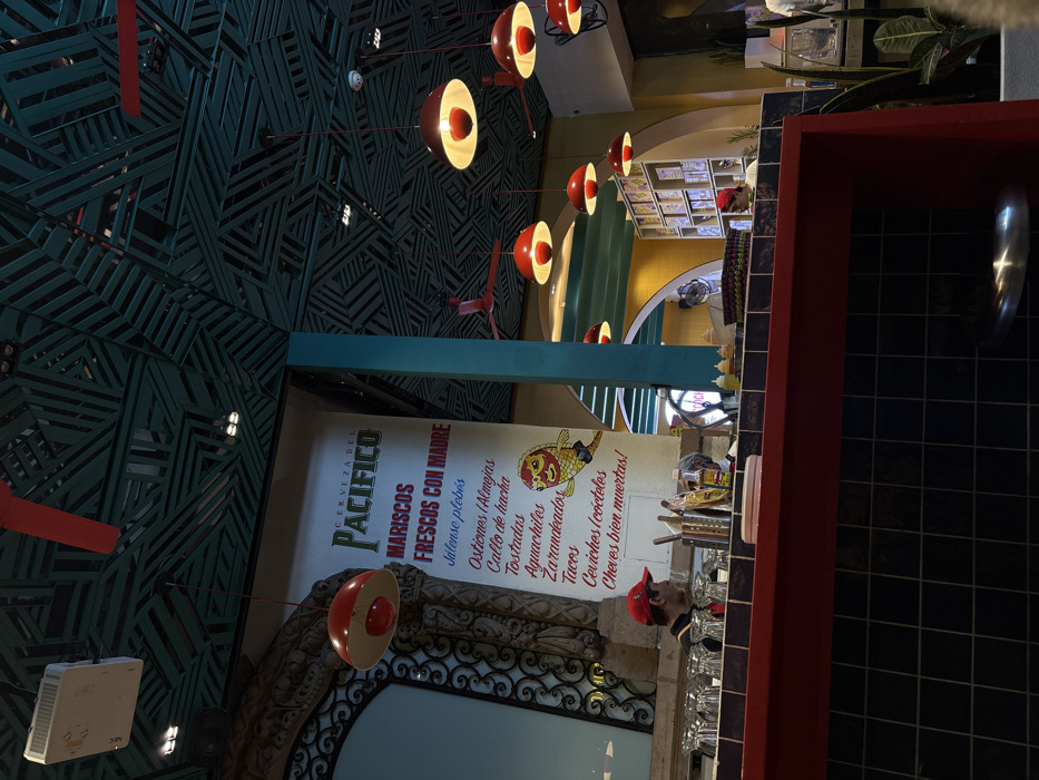 The image is an indoor shot of a restaurant featuring a menu board. The menu advertises "CERVEZA DEL PACIFICO" along with fresh seafood ("MARISCOS FRESCOS CON MADRE"). Food items listed include oysters, scallops, tostadas, aguachiles, grilled fish, tacos, ceviches, and cold beers ("Cheves bien muertas!"). A person wearing a red cap is visible near the bar area. The restaurant's décor includes a dark green patterned ceiling, artistic lighting fixtures, and tiled walls.
