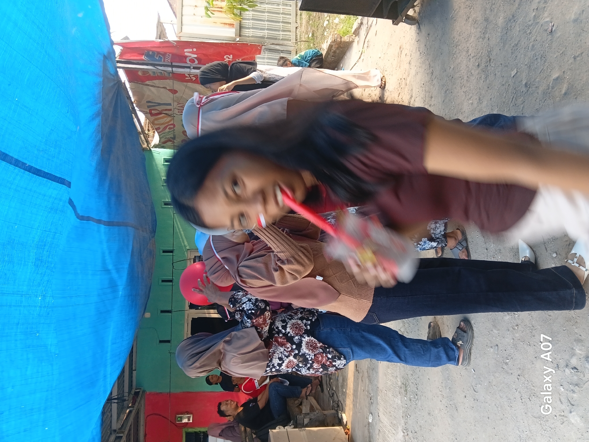 The image shows an outdoor scene, likely in Tembung, Indonesia, under a large blue tarp. In the foreground, a young woman with long dark hair is in motion, her face blurred and her body angled towards the viewer. She is holding a red and white striped cup with a red straw to her mouth, and appears to be drinking. A red stripe is visible on her cheek. Behind her, several other individuals are present. To the left, a person wearing a brown headscarf and a floral-patterned top with blue jeans is visible from the waist down. Further left, a man in a black t-shirt and jeans is sitting on a wooden bench, smiling. In the mid-ground, a person in a brown headscarf and sweater is holding a red balloon. Another person, also wearing a headscarf, is holding a red and white ribbon. The ground is dirt and gravel. In the background, buildings with colorful facades (red and green) and a white fence are visible. A red banner with text that is partially obscured hangs on the right side.