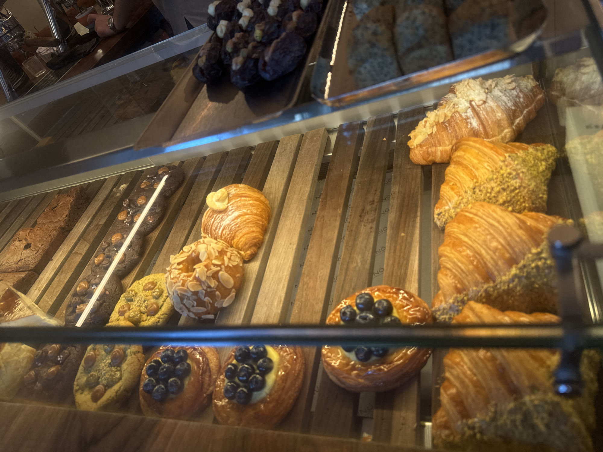 The image shows a display case filled with various baked goods, likely pastries and breads. The items include croissants, some plain and others with toppings like powdered sugar, almonds, pistachios, or a cream filling with blueberries. There are also cookies or other sweet breads with different toppings. The baked goods are arranged on wooden shelves. The image appears to be taken from inside a bakery or cafe. Given that the wooden shelves have the word "NO" on them, it is likely a bakery called "No" in Yerevan, Armenia. The angle of the photo suggests the viewer is either standing in front of or slightly above the display case. Parts of the cafe interior, including a person at a counter, are visible in the reflection on the glass of the display case. A metal bar obstructs the view in the foreground.