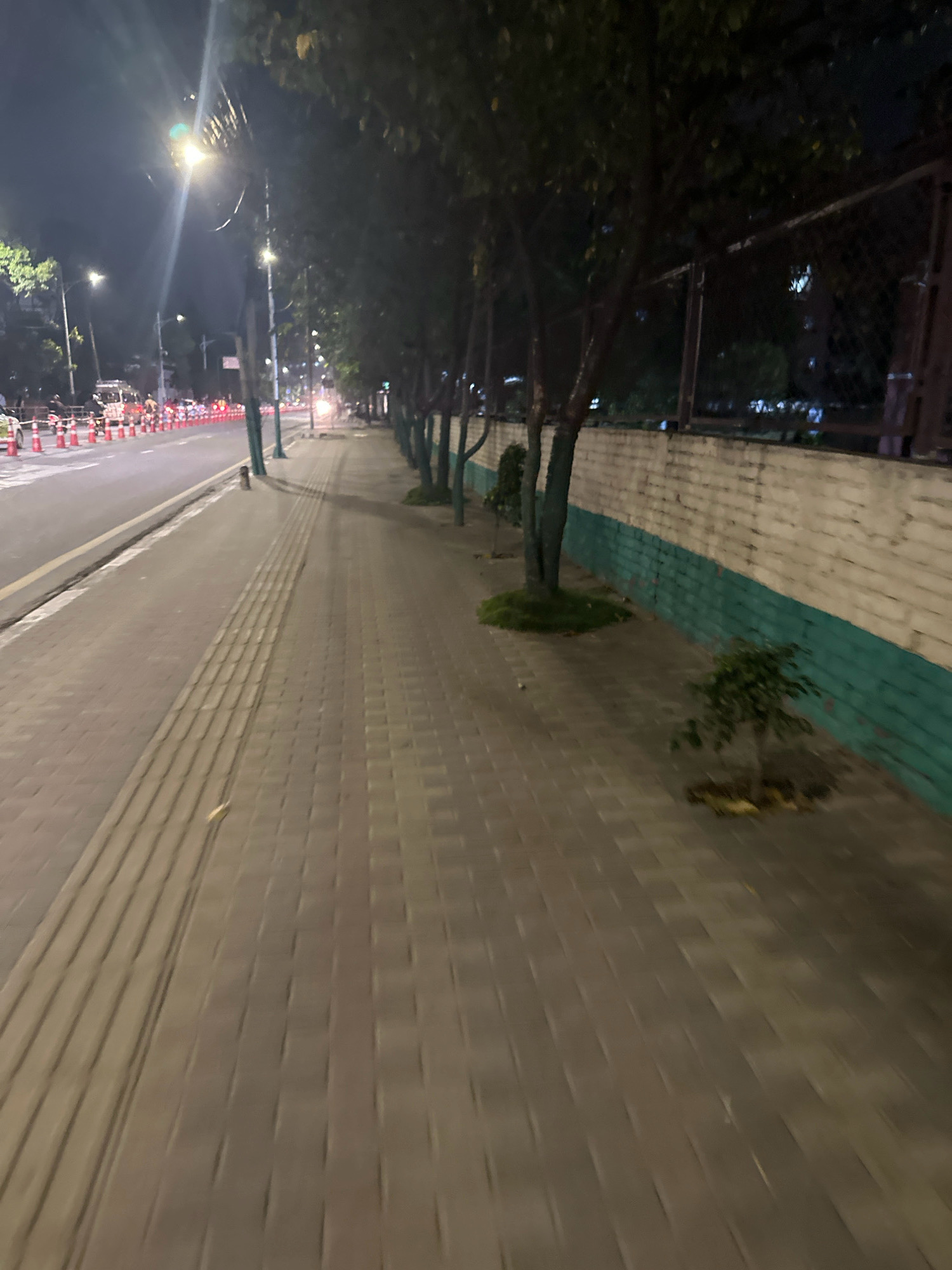 The image depicts a sidewalk at night in a city. The sidewalk is paved with rectangular bricks and has a tactile paving strip running along its length. A row of trees lines the sidewalk, with their branches casting shadows on the pavement. Beyond the trees, there is a low wall painted with a white upper half and a green lower half. A chain-link fence runs along the top of the wall. On the other side of the sidewalk is a street with traffic cones and what appear to be vehicles. Streetlights illuminate the scene. The overall impression is of an urban environment at night, possibly in a residential or commercial area. Without specific landmarks or identifying features, it is not possible to determine the precise location within Kathmandu Metropolitan City, Nepal.