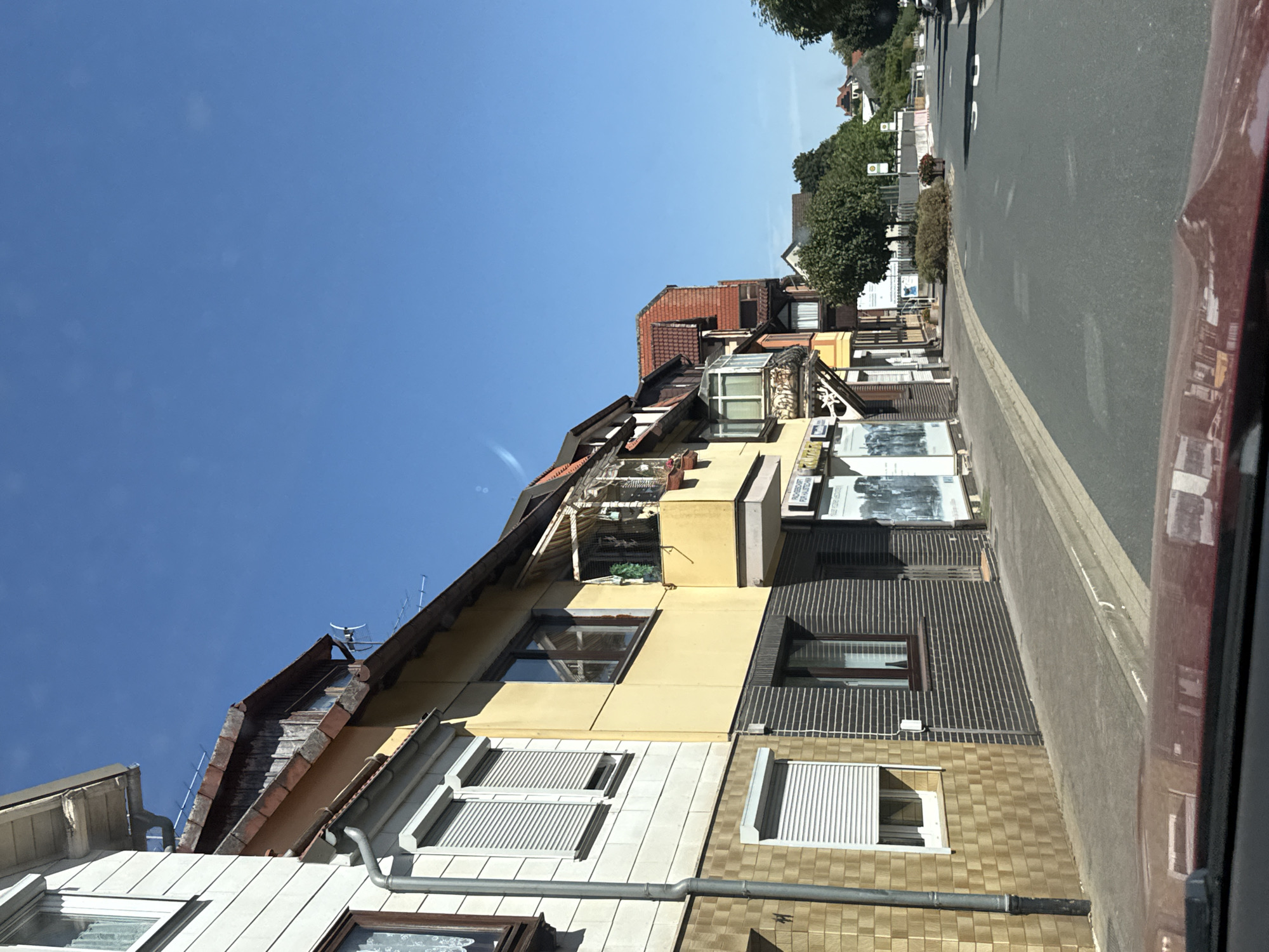 This is an eye-level, medium shot of a street in the town of Bad Sachsa, Germany, on a clear, sunny day. The view is from inside a vehicle, as indicated by the red dashboard in the foreground and the reflection of the buildings on it.
On the left side of the street, a row of two-story buildings with businesses on the ground floor is visible. The building closest to the viewer has a light-colored facade with white horizontal siding and a lower section covered in tan square tiles. It features windows with white roller shutters and a prominent grey downspout running vertically along its side. The building next to it is painted yellow and has dark brick detailing on its lower level. It has a window with a brown frame and a balcony with an awning above it, decorated with plants. Further down the street, other buildings with red-tiled roofs are visible, some with balconies and awnings. One building has a sign that reads "FACHGESCHÄFT FÜR HAUSSTECHNIK".
The street itself is paved and asphalted, with visible lane markings and some white painted arrows indicating traffic direction. A sidewalk runs alongside the buildings. Green trees and bushes are present on the right side of the street, as well as in the background, leading towards more houses with red roofs and trees. The sky is a clear, bright blue with no clouds. There are no people visible in the image. The scene appears to be a residential and commercial street in a German town during daylight hours.