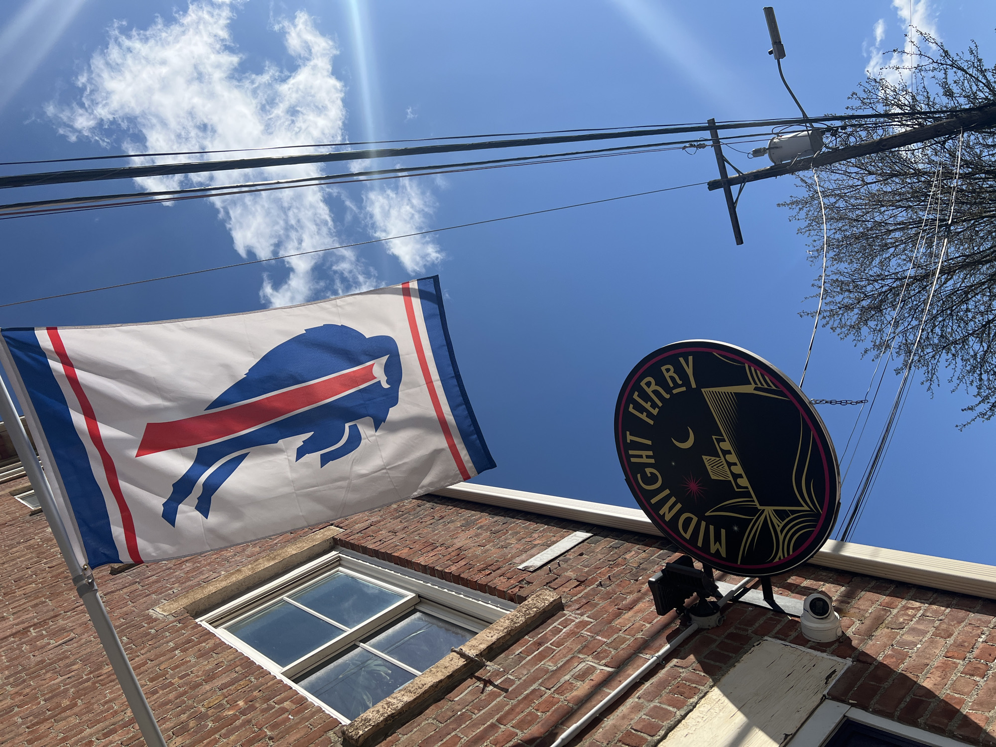 The photograph, taken in the City of Newburgh, United States of America, depicts the exterior of a brick building under a bright, blue sky with scattered clouds. Visible on the left is a white flag bearing a blue and red Buffalo emblem, a symbol often associated with the Buffalo Bills football team. To the right, an oval-shaped sign with the words "NIGHT FERRY MIDNIGHT" in a circular pattern encircles a depiction of a ship with a crescent moon above it, indicating the name of a possible establishment. A surveillance camera is mounted below the sign. Above, power lines crisscross the sky, connecting to a utility pole with electrical equipment and bare tree branches extending towards the upper-right corner of the frame. A window with a white frame is visible on the brick facade.