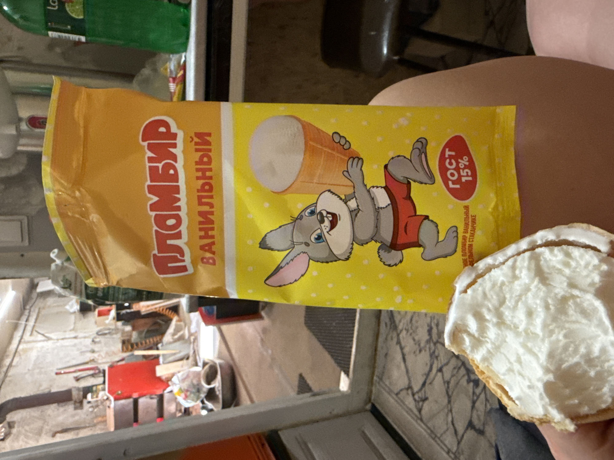 A close-up, indoor shot shows a partially eaten ice cream sandwich and a package of Plombir ice cream in front of a person. The package is yellow with a cartoon rabbit holding an ice cream cone, and the text "ПЛОМБИР ВАНИЛЬНЫЙ" (Plombir Vanilla) and "ГОСТ 15%" is visible. The ice cream sandwich is held in the person's hand. The background reveals a cluttered interior, with a view through a window showing a messy room or workshop. The objects, actions, and interactions suggest a casual, domestic setting. It is difficult to ascertain the location in the city of Chaikovsky, Russia. The setting could be a home, workshop, or similar indoor space.