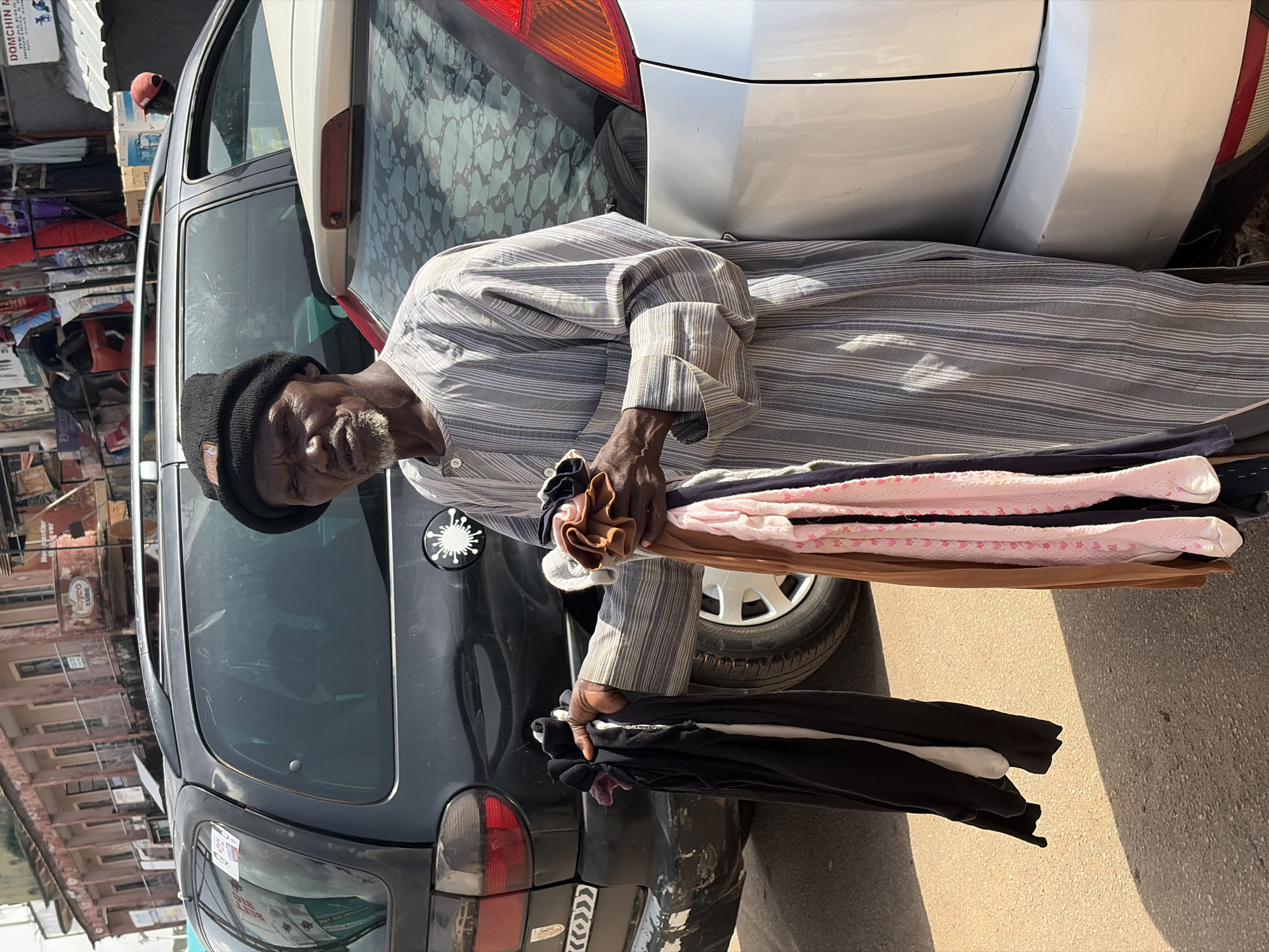 An elderly Black man wearing a dark beanie and a grey and white striped robe stands in front of a silver car, holding a bundle of folded clothes. The clothes include a black garment, a white garment, a brown garment, and pink garments with a heart pattern. Behind him is a black vehicle and buildings. The scene appears to be a street in Jos, Nigeria. The man's expression is stern. The lighting suggests it is daytime, with strong shadows indicating direct sunlight.