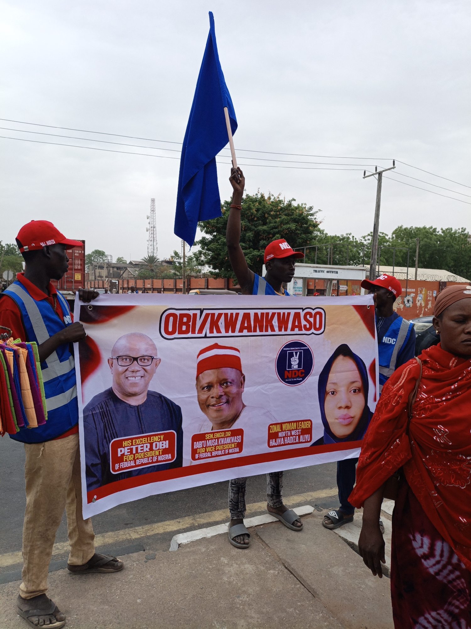A group of people are holding a banner for the political candidates Obi/Kwankwaso in an urban area. The banner features photos of Peter Obi, Rabi'u Musa Kwankwaso, and Hajiya Hadiza Aliyu, with text indicating their positions in the Nigerian government. A man in the center holds a blue flag. The individuals on either side of the banner wear red caps and blue vests, one holding colorful textiles. A woman in a red dress walks in front of the banner. In the background, there are buildings, a traffic officer post, shipping containers, and an antenna tower against an overcast sky. The location appears to be a street or sidewalk.