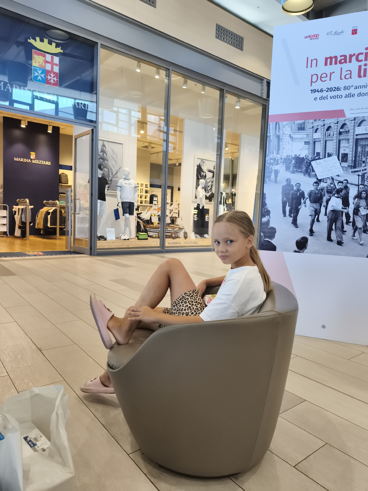 A young girl with blonde hair tied in a ponytail sits in a gray armchair in an indoor commercial space. She is wearing a white t-shirt and a leopard-print skirt. Her feet are resting on the seat of the chair, and she is wearing pink slippers. To her left, a white plastic bag is visible on the floor. Behind her, a large poster displays a black and white historical image and text in Italian. To her right, the storefront of "MARINA MILITARE" is visible, featuring mannequins dressed in clothing. The floor is tiled with light-colored wood-effect tiles. The environment appears to be a mall or shopping center. There is no visible indication of the city of Livorno, Italy in the image.