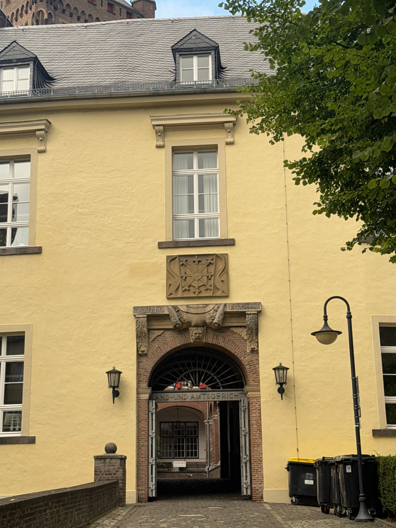 The image shows the exterior of a yellow, two-story building with an arched entranceway. Above the entrance, there is a stone relief with a crest and text that appears to say "ANNO 1664." The building is located in the city of Cleves, Germany, and appears to be part of a historical complex. To the right of the entrance, there are three trash bins. A lamppost is situated next to the bins, and a large tree with green leaves overhangs the building from the right. The sky is visible in the background, with a cloudy overcast. A cobblestone pathway leads up to the entrance, and a brick wall is visible to the left. The overall impression is of an old, well-maintained building with historical significance.