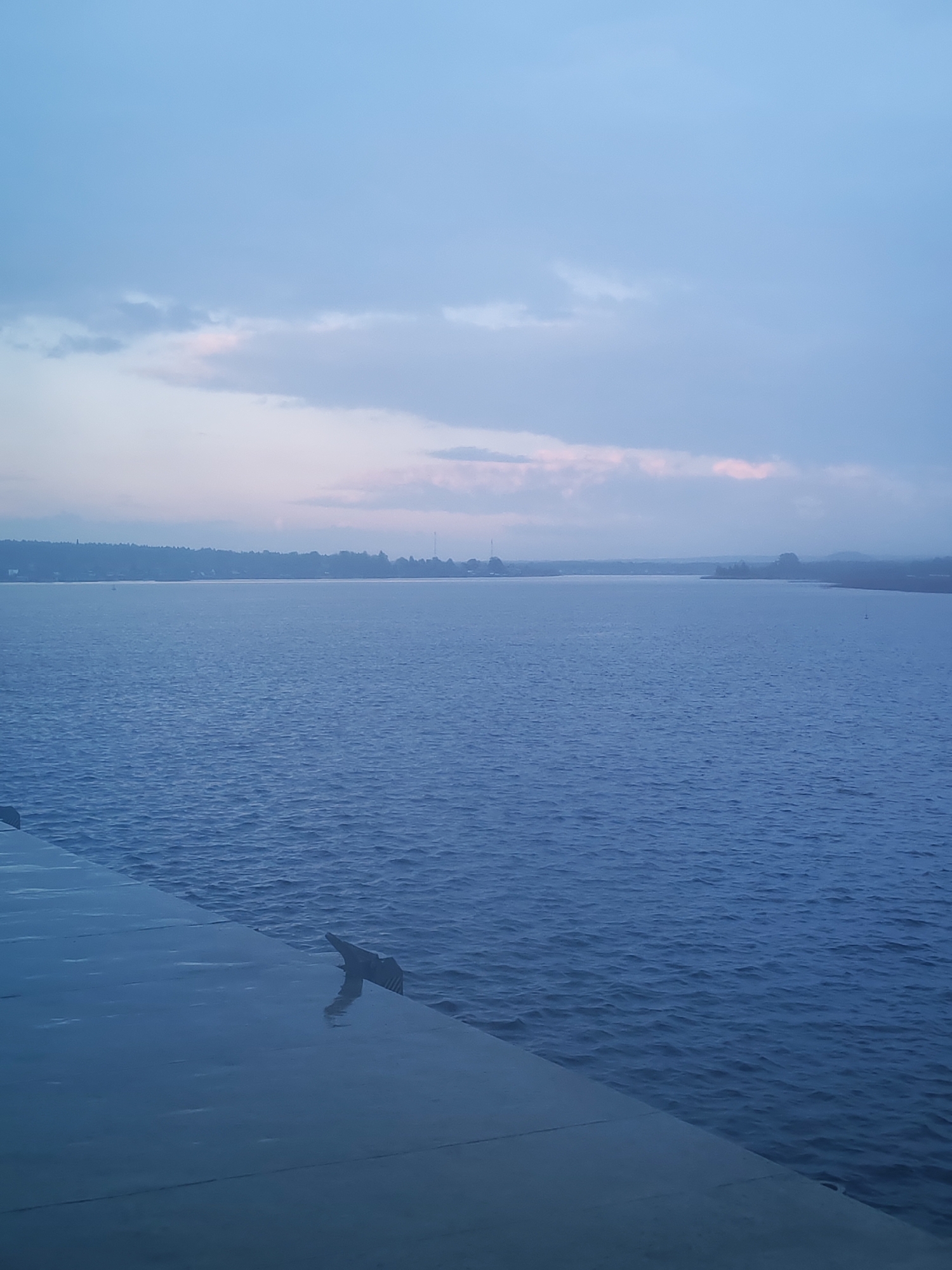The image shows a body of water, likely a river or bay, under a cloudy sky with hints of pink near the horizon. In the foreground, a wet, reflective surface suggests a quay or dock area. The water surface shows ripples and small waves. Across the water, a distant shoreline is visible with trees and some structures, potentially a part of the city of Shalsky, Russia. The lighting is dim and subdued, indicating either dusk or a heavily overcast day. A dark, protruding object is present on the quay, possibly a bollard or mooring fixture.