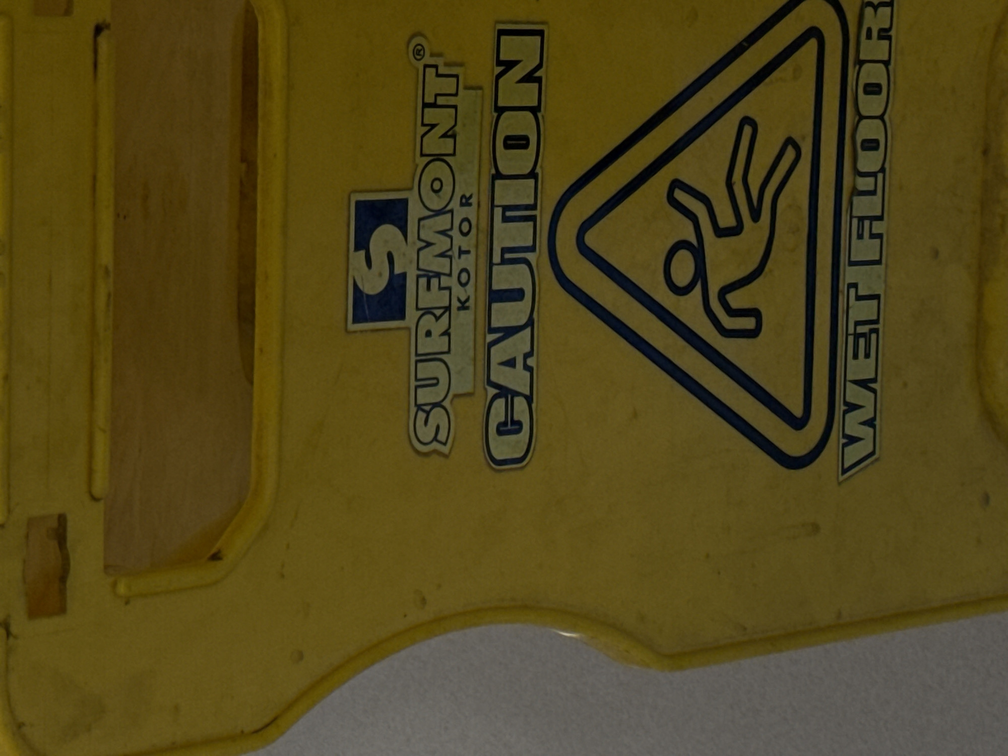 The image presents a close-up, slightly rotated view of a yellow plastic "Wet Floor" warning sign. The sign is positioned vertically, with its longer dimension extending from the top to the bottom of the frame. The background is a plain, neutral-toned surface, possibly a wall, transitioning to a lighter grey area at the bottom right, which could be the floor the sign rests upon.
The sign features multiple texts and a graphic, primarily in blue and white. On the left side, there's a square blue logo with a white, stylized "S." To its right, the word "SURFMONT" is printed in large, blue-outlined capital letters, accompanied by a registered trademark symbol (®). Below "SURFMONT," the word "KOTOR" is visible in smaller blue-outlined capitals.
Dominating the center of the sign is the word "CAUTION," printed vertically in large, bold, blue-outlined capital letters. Adjacent to this, a prominent triangular warning symbol is displayed. This symbol has a thick blue border and contains a blue silhouette of a person in mid-fall, arms and legs extended, indicating a slipping hazard.
On the far right edge of the sign, the phrase "WET FLOOR" is printed vertically in bold, blue-outlined capital letters. The overall appearance suggests a functional safety sign used in an indoor environment to warn about slippery surfaces. There are no people visible, and no active event is taking place other than the implied warning for a wet floor condition. The sign shows some signs of use and minor wear. The time of day and weather are not discernible.