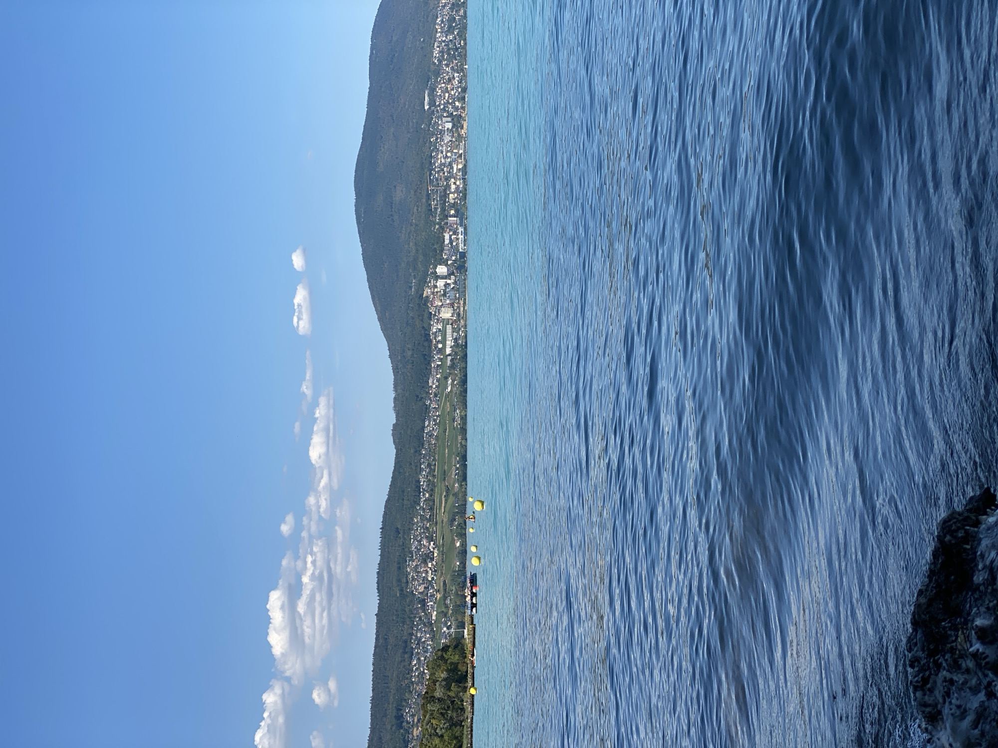 A wide, high-angle shot captures a tranquil scene of a body of water with a distant, built-up shoreline and a verdant hill under a clear blue sky. In the foreground, dark, wet rocks are visible at the bottom left, transitioning into a rippling blue expanse of water that takes up the majority of the frame. The water transitions to a lighter, turquoise hue in the middle ground, where several yellow buoys are scattered. A small, dark, floating structure, possibly a raft or dock, is situated amongst the buoys, with a few indistinct figures and small boats nearby. The background features a densely populated town nestled at the foot of a rolling, tree-covered hill. The architecture of the town includes numerous white and light-colored buildings, some with visible cranes, suggesting ongoing construction. The hill behind the town is dotted with more trees and appears to have terraced vineyards on its slopes. The sky above is a bright, cloudless blue, with a few scattered white clouds near the horizon. This image was likely taken from a location near the shore of Lake Neuchâtel in Cortaillod, Switzerland.