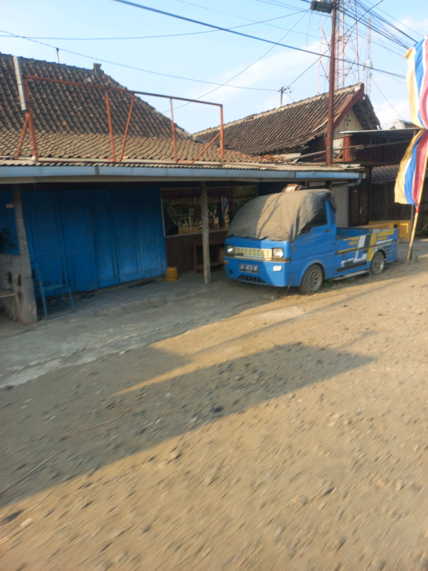 A blue truck, partially covered by a grey tarp, is parked on a dirt road in front of a building with blue shutters in Baturetno, Indonesia. The building has a tiled roof with exposed wooden beams. Overhead, power lines crisscross the sky, and a cell tower is visible in the distance. A few colorful flags are attached to a pole on the right side of the frame. The ground is dusty and uneven, with shadows cast by the building and the truck.
