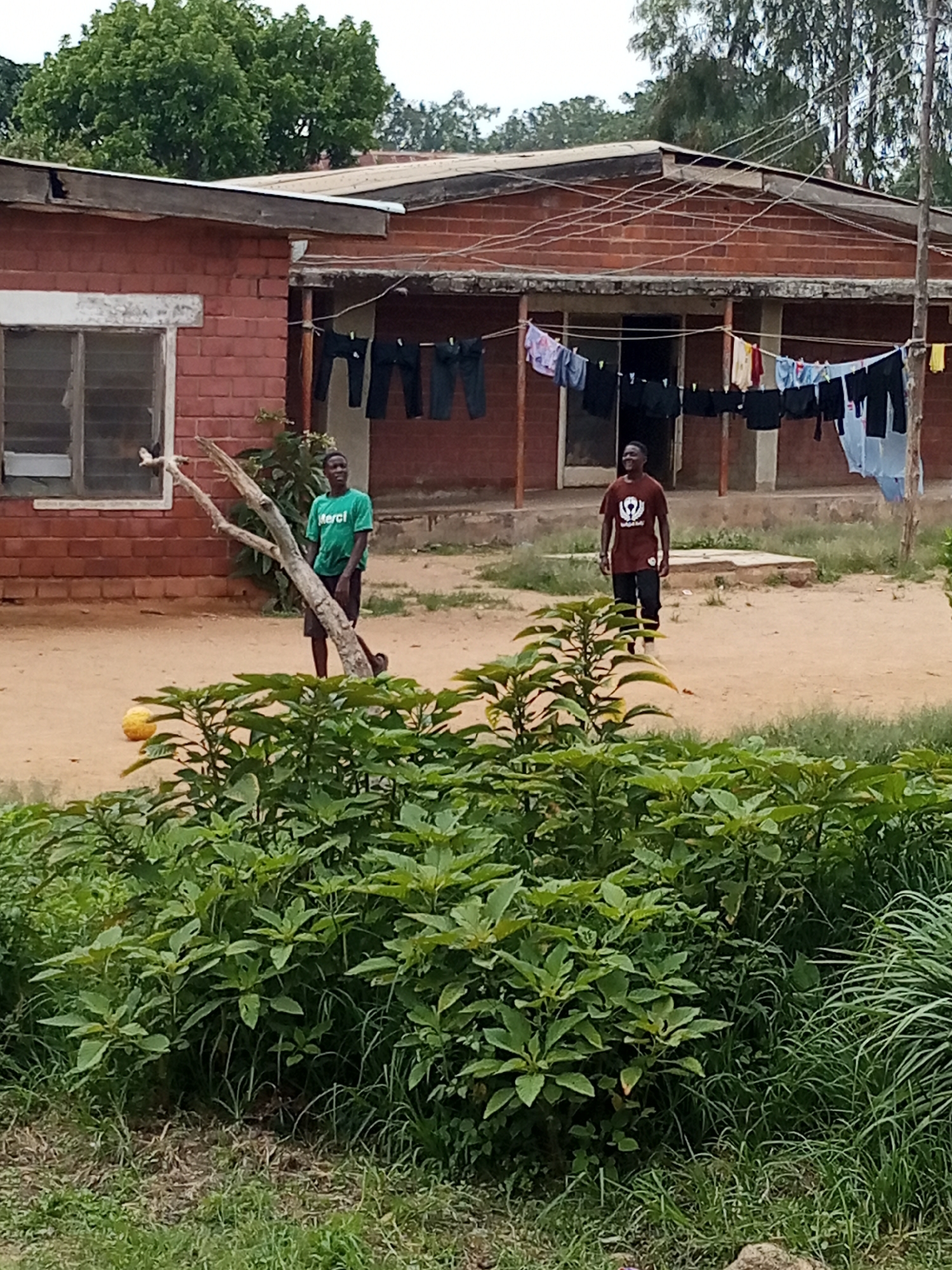 In Jos, Nigeria, an outdoor scene depicts two young men in front of a brick building with a corrugated iron roof. Laundry hangs on a clothesline strung across the front of the building. The ground is dirt, with some patchy grass and greenery. One man, wearing a green t-shirt with the word "Merci" on it and dark shorts, stands near a dead tree trunk. To his right, another man, in a maroon t-shirt and dark pants, stands with his hands in his pockets, looking to the side. A yellow, ball-like object rests on the ground to the left of the first man. The building has a window with a broken pane on the left. The sky is overcast.