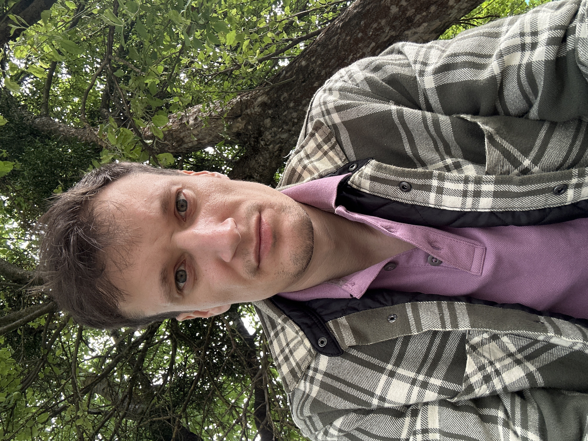 A medium shot selfie is taken from a low angle, looking up at a man sitting outdoors. The man has fair skin, light brown hair, and light blue eyes. He is wearing a pink collared shirt and a green and white plaid jacket over a black quilted vest. He is looking directly at the camera with a neutral expression. Behind the man, a large tree trunk is visible, with branches and green leaves forming a canopy overhead. The lighting suggests it is daytime, possibly overcast. The setting appears to be a park or natural area. The image's origin is not specified, but it is presented in the context of Makiv, Ukraine.