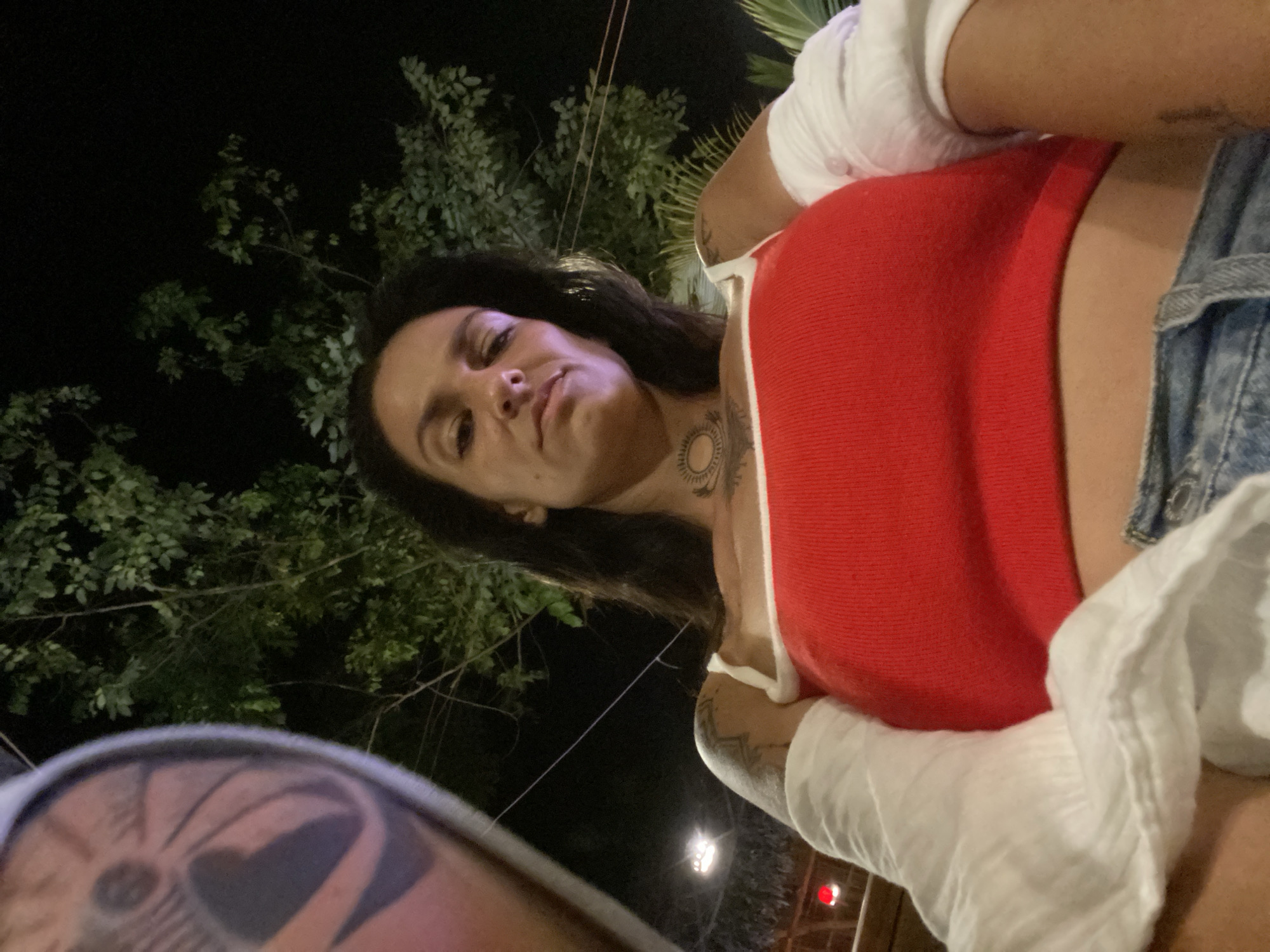 The image shows a woman in a red top and denim shorts, with her arm resting on a white, textured garment. She has tattoos on her arms and neck. The background features green foliage against a dark, night sky, with some distant lights that could indicate an outdoor setting in a city. The camera is positioned low, looking up at the woman, suggesting a selfie or a photo taken from a low angle. There is a blurry tattoo on the left side of the frame, possibly belonging to another person. The presence of outdoor lighting and greenery could suggest a restaurant, bar, or park setting in a tropical or subtropical urban area.