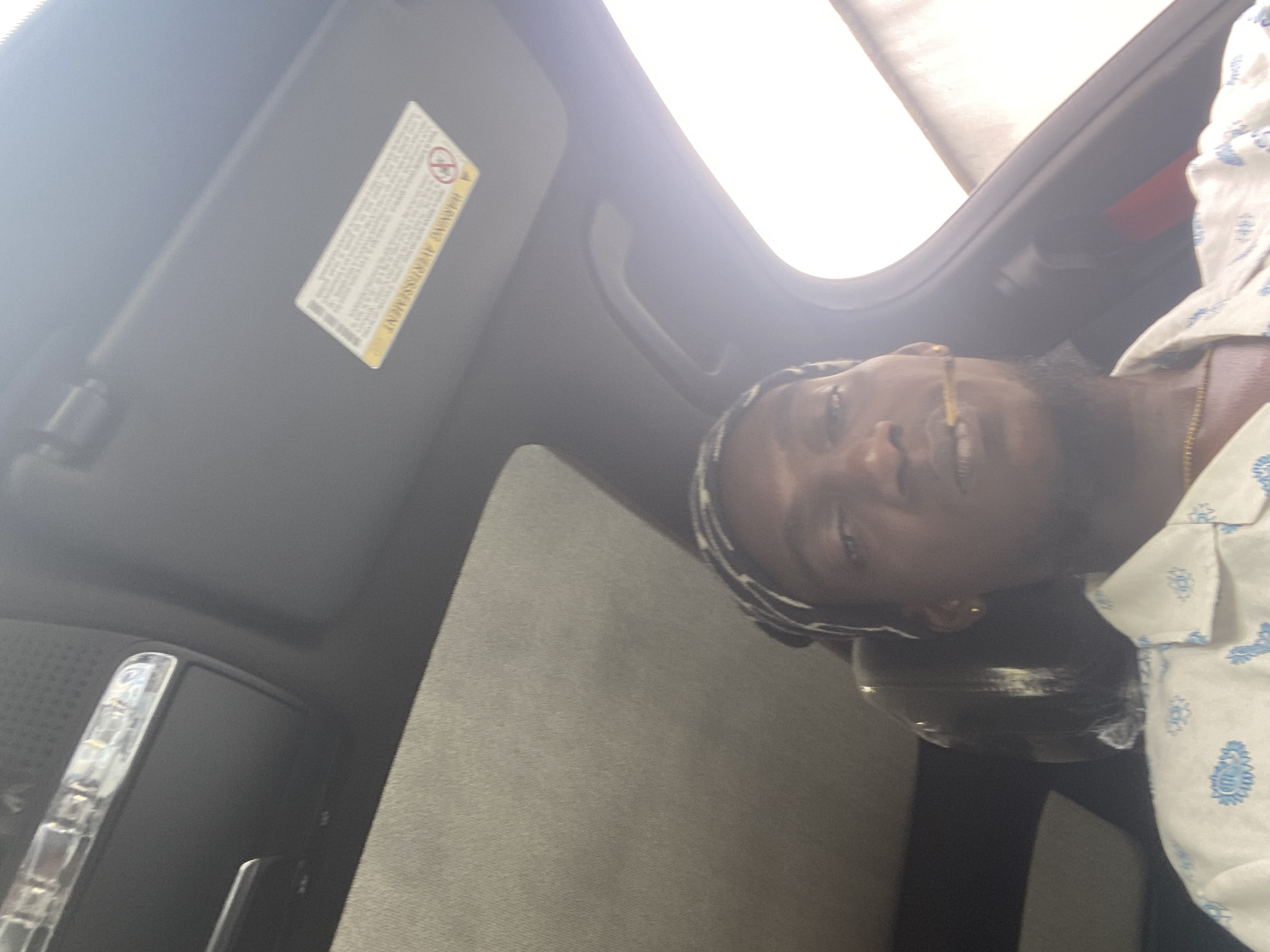 A man is seen from the chest up, inside a car. He is wearing a white shirt with a blue pattern and a bandana on his head. He has a cigarette in his mouth. The car's sunroof is open, and the sun is shining brightly. A warning label is visible on the car's visor. The image appears to be taken in Oniyangi, Nigeria, as specified in the prompt.