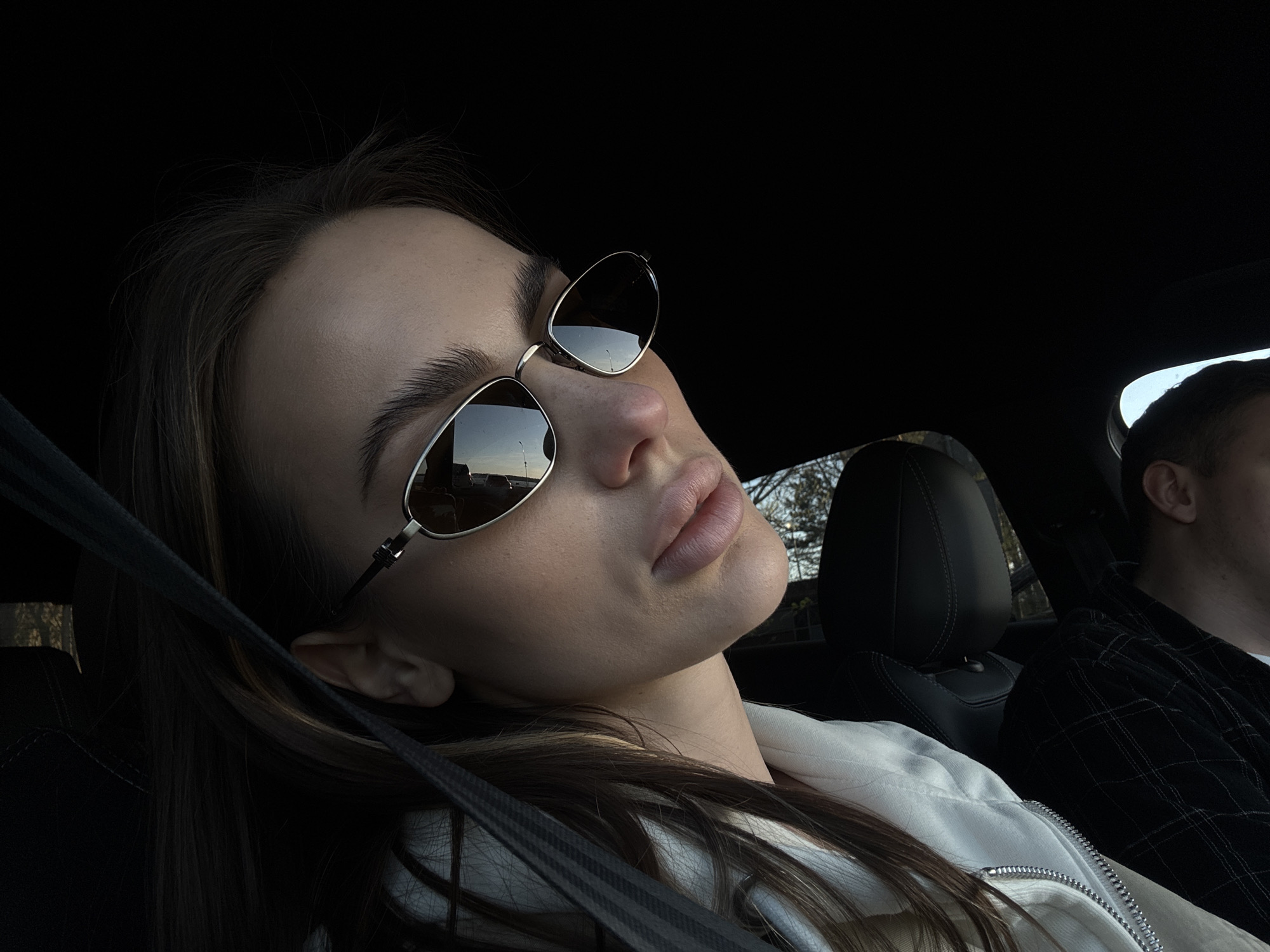 The image captures an intimate interior view of a car, likely in Białobrzegi, Poland, during what appears to be late afternoon or early evening due to the soft, warm lighting.
The main subject is a young woman, positioned as a passenger in the front seat, leaning her head back against the black headrest. She is seen from the chest up, wearing fashionable dark sunglasses with highly reflective lenses. The reflections in her sunglasses clearly show an outdoor scene, including a road or parking area, some distant structures or trees, and a bright, clear sky, indicating fair weather. Her dark, long hair is visible, flowing around her head and shoulder. She is wearing a light-colored, possibly white, zippered jacket, and a dark car seatbelt runs diagonally across her chest and shoulder. Her lips are slightly parted, and her overall expression appears relaxed or thoughtful, possibly posing for the photograph.
To the right side of the frame, the top of a man's head and his shoulder are partially visible, suggesting he is in the driver's seat. He is wearing a dark, possibly plaid, shirt. The car's interior is predominantly dark, with black upholstery and headrests. The lighting casts soft shadows, highlighting the woman's face and the details of her sunglasses and jacket. There is no visible text in the image. The scene conveys a moment of calm or travel within the vehicle.
