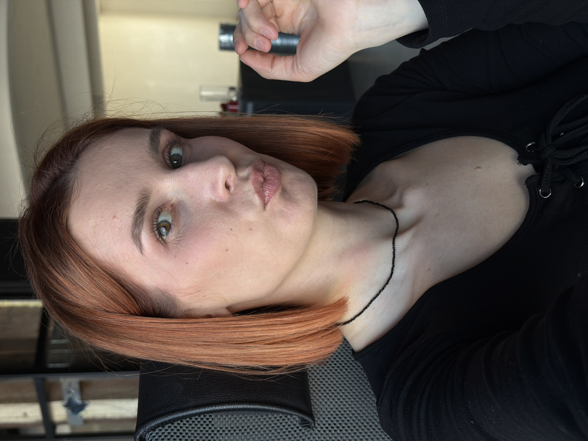 A woman with auburn hair is posing for a selfie. She is wearing a black top with a laced neckline and a thin black necklace. Her lips are puckered. The background is blurred, but appears to show a window, desk, and chair. There is no information indicating the location of the image to be in Chaikovsky, Russia.