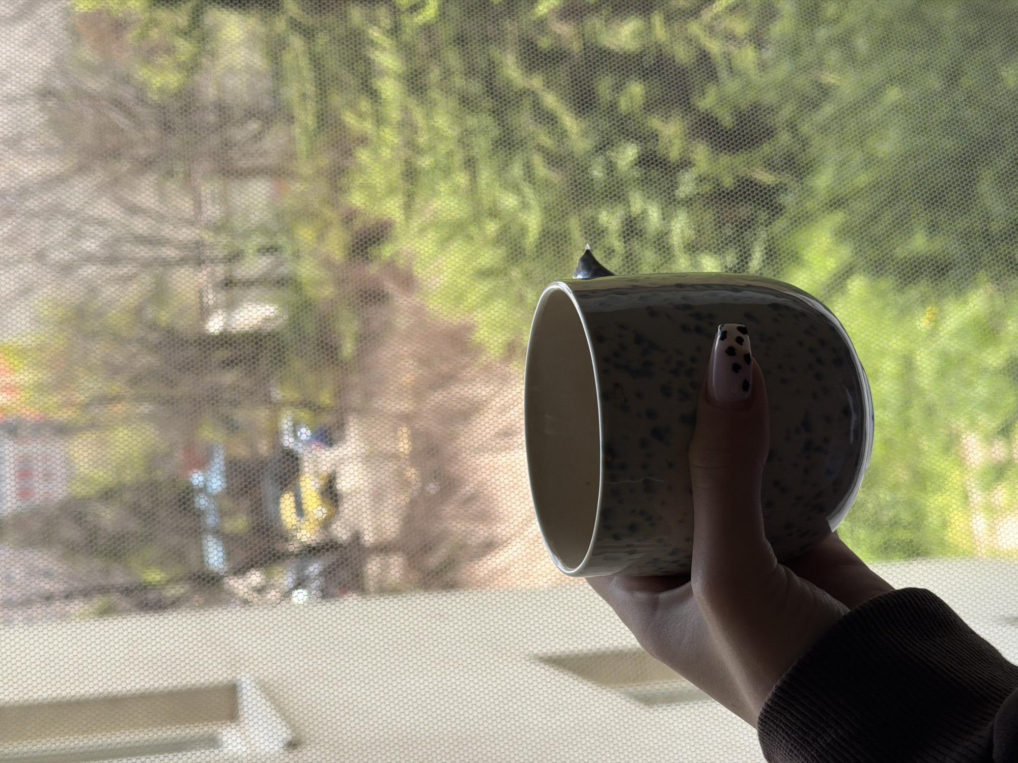A close-up image captures a person's hand holding a ceramic mug against a blurred outdoor background, viewed through a fine hexagonal mesh screen. The hand, positioned on the right side of the frame, is wearing a dark, possibly brown, long-sleeved garment. One of the person's fingernails features a distinct black and white spotted pattern. The mug is light-colored with dark speckles, and a small, dark, pointed design element protrudes from its rim. It appears to be mostly empty.
The background, heavily filtered by the mesh, shows a blend of vibrant green foliage and the starker, bare branches of trees, suggesting either a transitional season like early spring or autumn, or a mix of evergreen and deciduous trees. Distant, indistinct shapes of buildings or structures are also visible through the haze. The scene is illuminated by natural light, indicating it is daytime. No specific activity beyond holding the mug is depicted, and no emotions are discernible. No text is visible in the image. The location context provided is Sofia, Bulgaria.