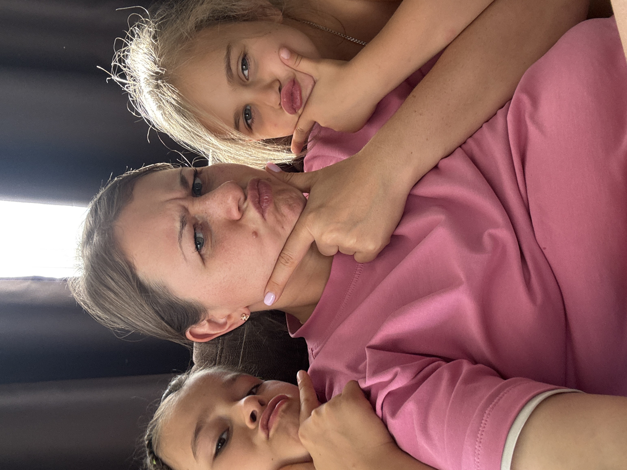 In the city of Uyutnoye, Ukraine, three individuals are positioned closely together, likely a mother and her two daughters, as indicated by their shared facial features and familial closeness. They are seated, with the mother at the center and the daughters on either side, in what appears to be a living room or personal space. The backdrop consists of a dark curtain and a window, suggesting an indoor setting. The overall ambiance is relaxed and playful, with all three subjects making exaggerated facial expressions, puckering their lips and pointing to their chins. The mother wears a pink t-shirt, and her daughters are also casually dressed. The lighting appears natural, casting soft shadows and highlighting their features. The image captures a moment of lighthearted interaction, showcasing a close familial bond.